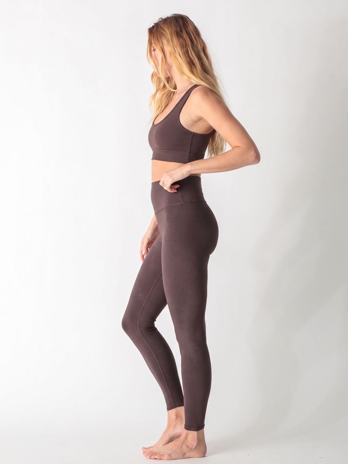A woman wearing a dark brown scoop neck sports bra and matching high-waisted leggings against a white background.
