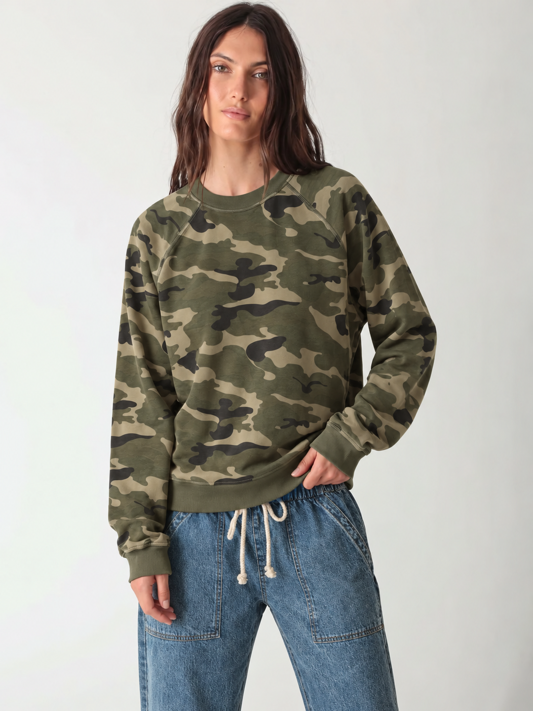 Model in an oversized green and khaki camo raglan crewneck sweatshirt with ribbed trim, half-tucked into light-wash drawstring denim pants.