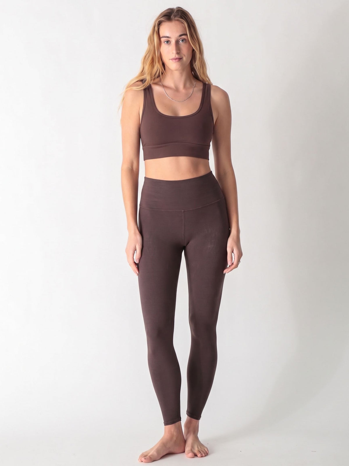 A woman wearing a dark brown scoop neck sports bra and matching high-waisted leggings against a white background.