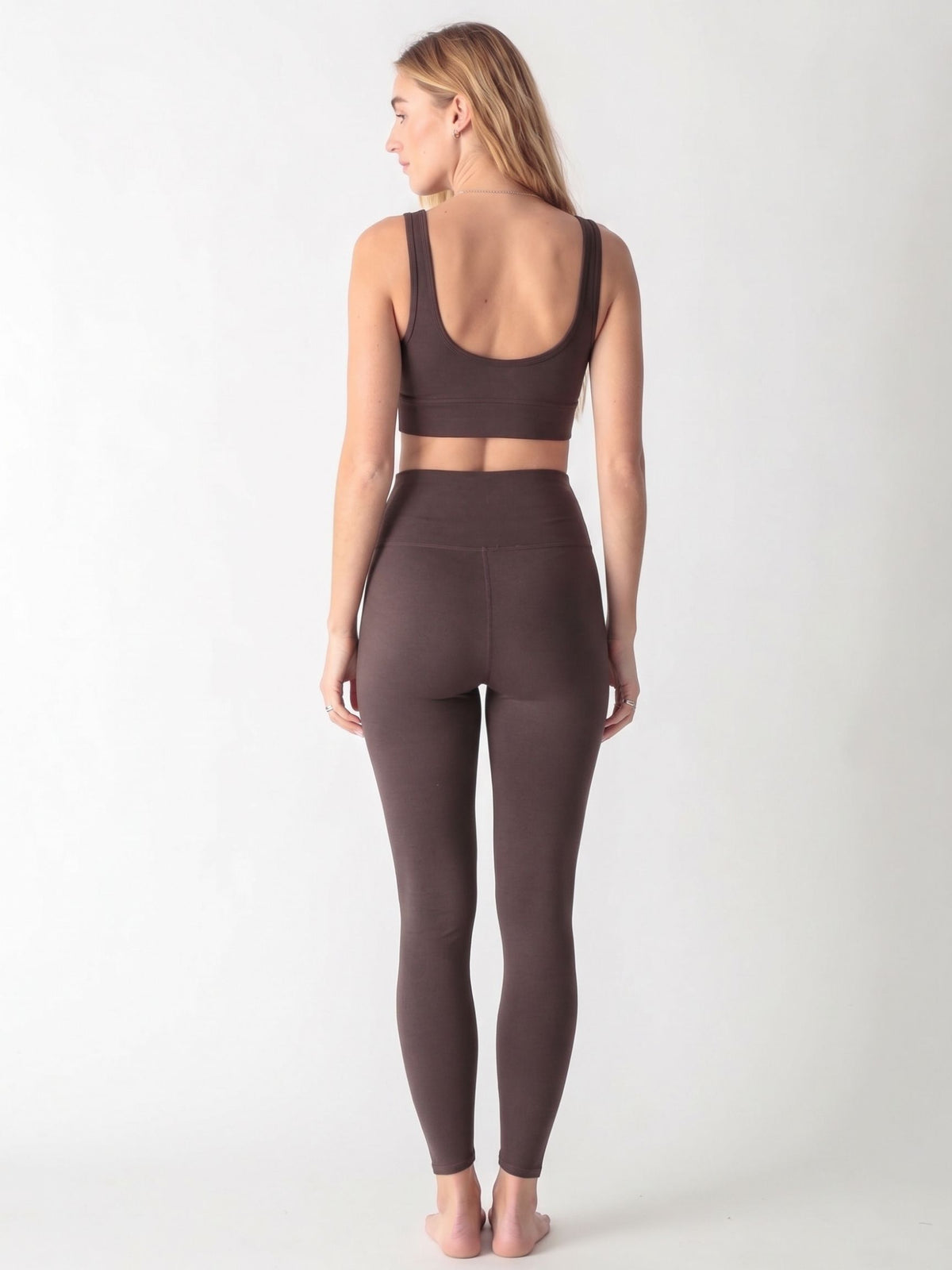 A woman wearing a dark brown scoop neck sports bra and matching high-waisted leggings against a white background.