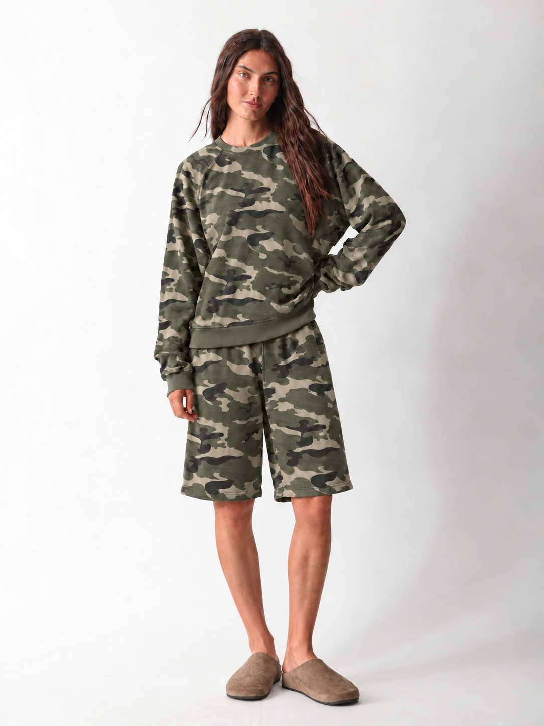 Model wearing a matching green camo set: a relaxed raglan-sleeve crewneck sweatshirt with ribbed cuffs and hem, and knee-length drawstring sweat shorts, styled with taupe suede clogs.