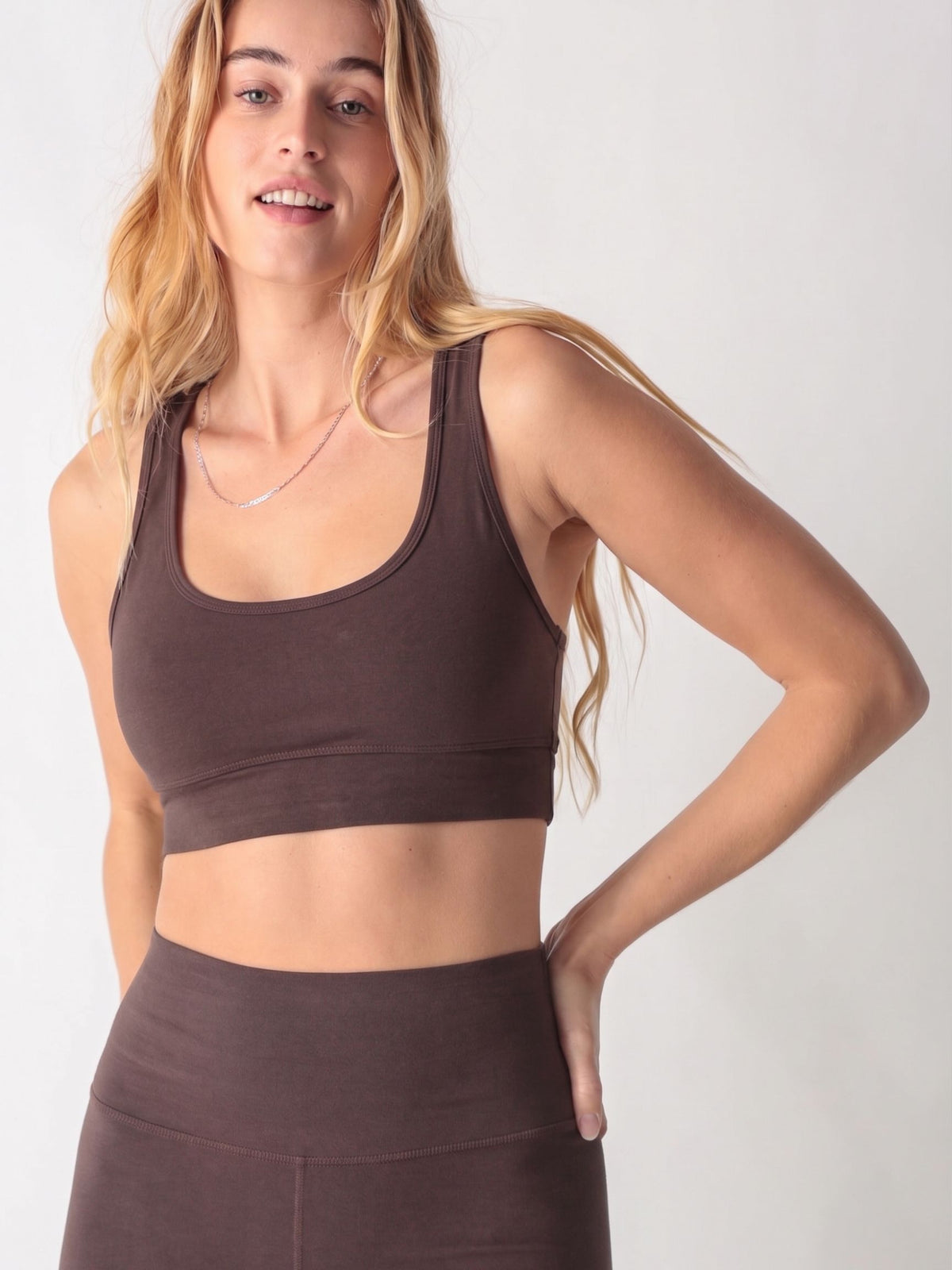 A woman wearing a dark brown scoop neck sports bra and matching high-waisted leggings against a white background.