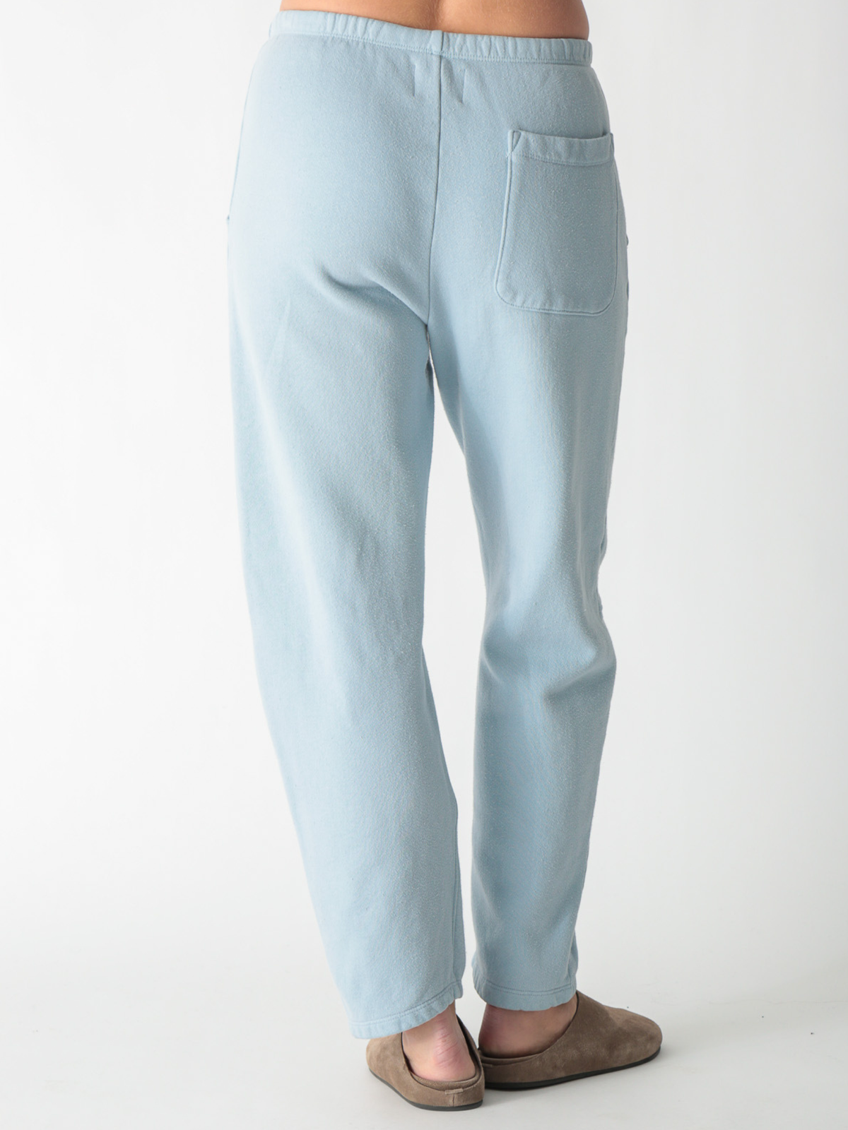A model wearing ice blue barrel-fit fleece sweatpants with elastic waistband, side pockets, and knee seams, paired with tan suede slip-on shoes against a plain white background.