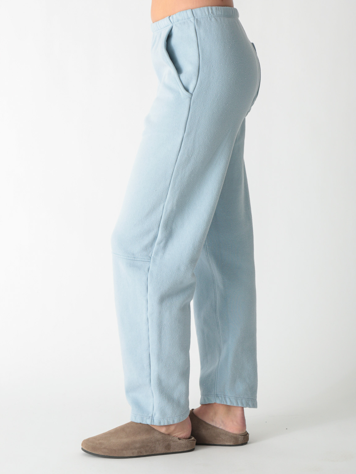 A model wearing ice blue barrel-fit fleece sweatpants with elastic waistband, side pockets, and knee seams, paired with tan suede slip-on shoes against a plain white background.