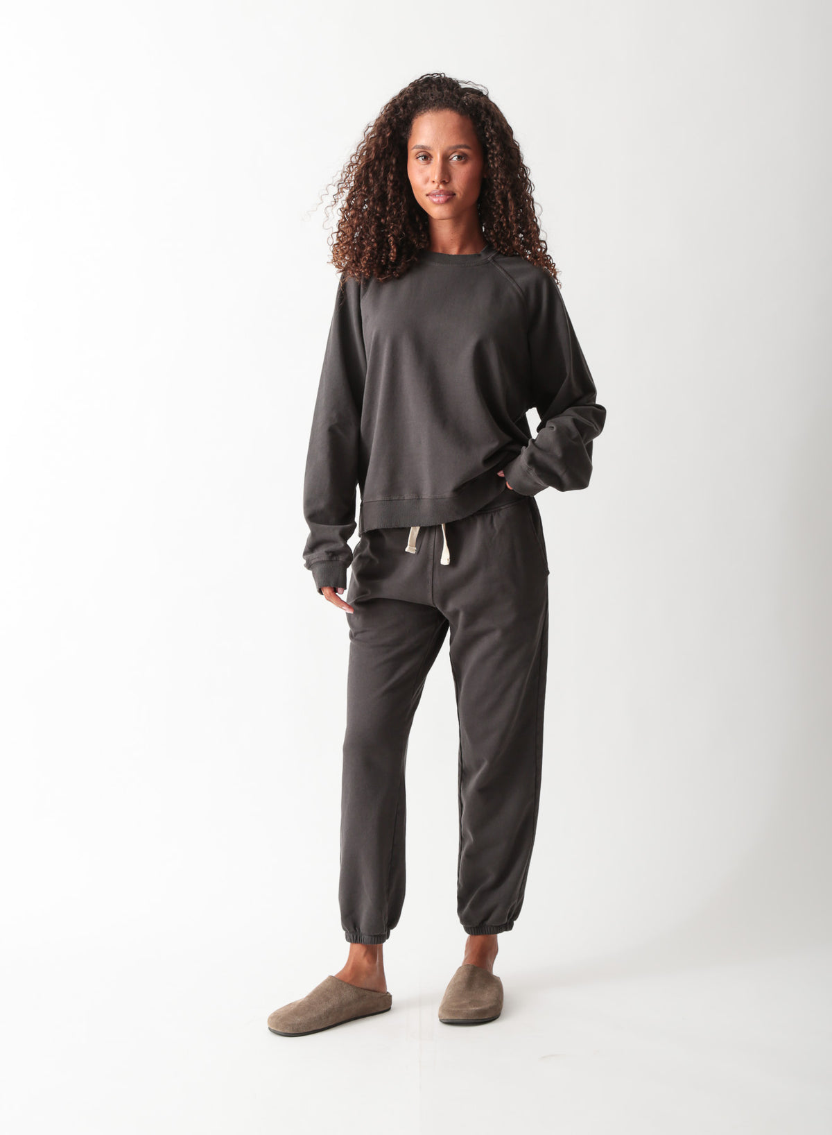 Woman wearing a washed charcoal raglan crewneck sweatshirt with matching drawstring jogger sweatpants and suede clogs.