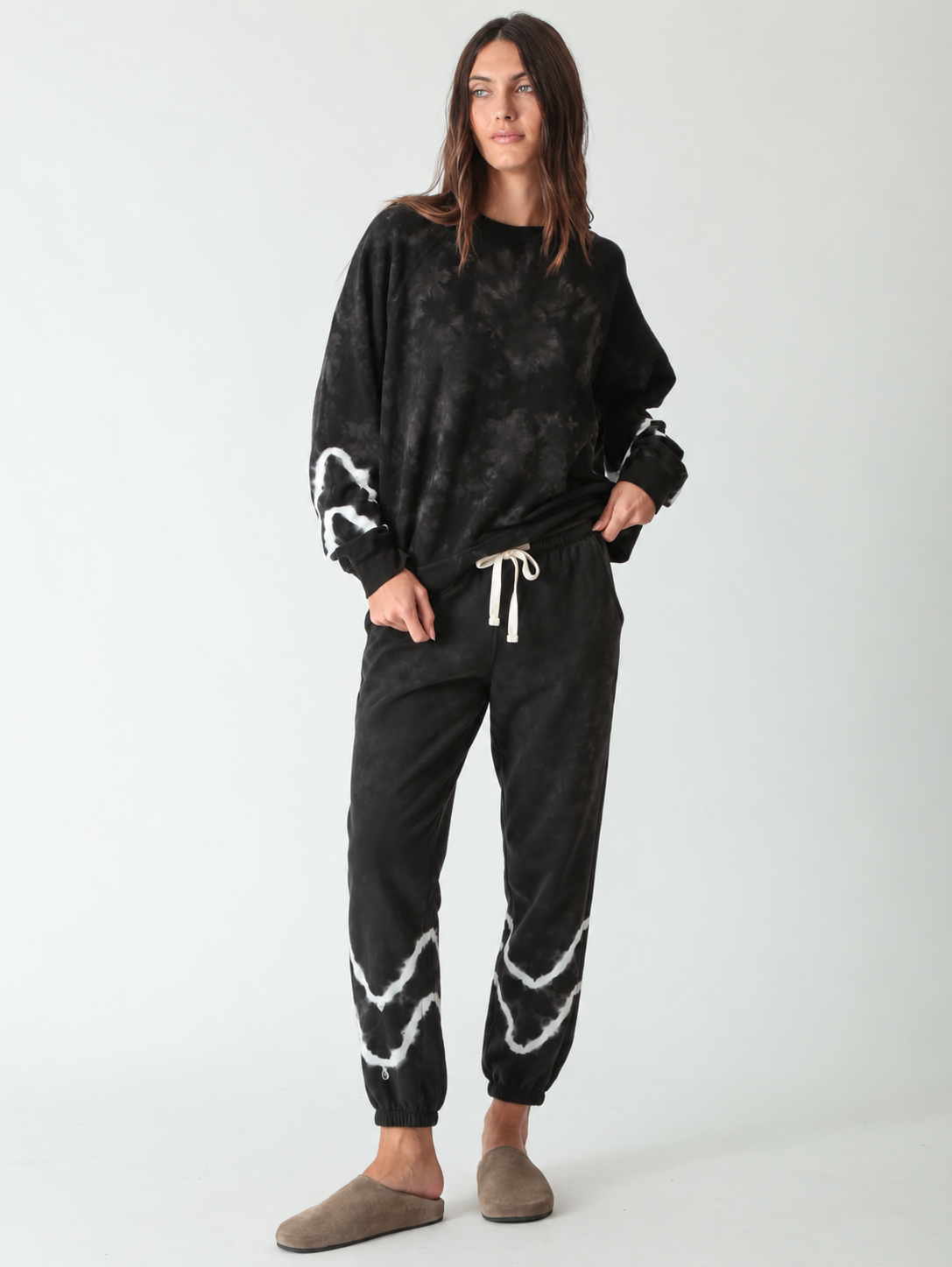 Woman wearing a black tie-dye crewneck sweatshirt with matching black jogger sweatpants with white wave detail and taupe suede clogs