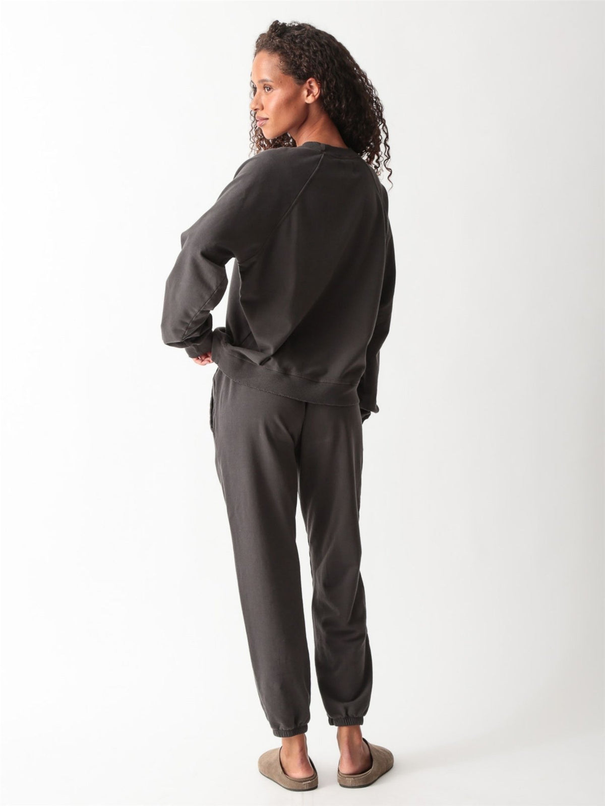 Woman wearing a washed charcoal raglan crewneck sweatshirt with matching drawstring jogger sweatpants and suede clogs.