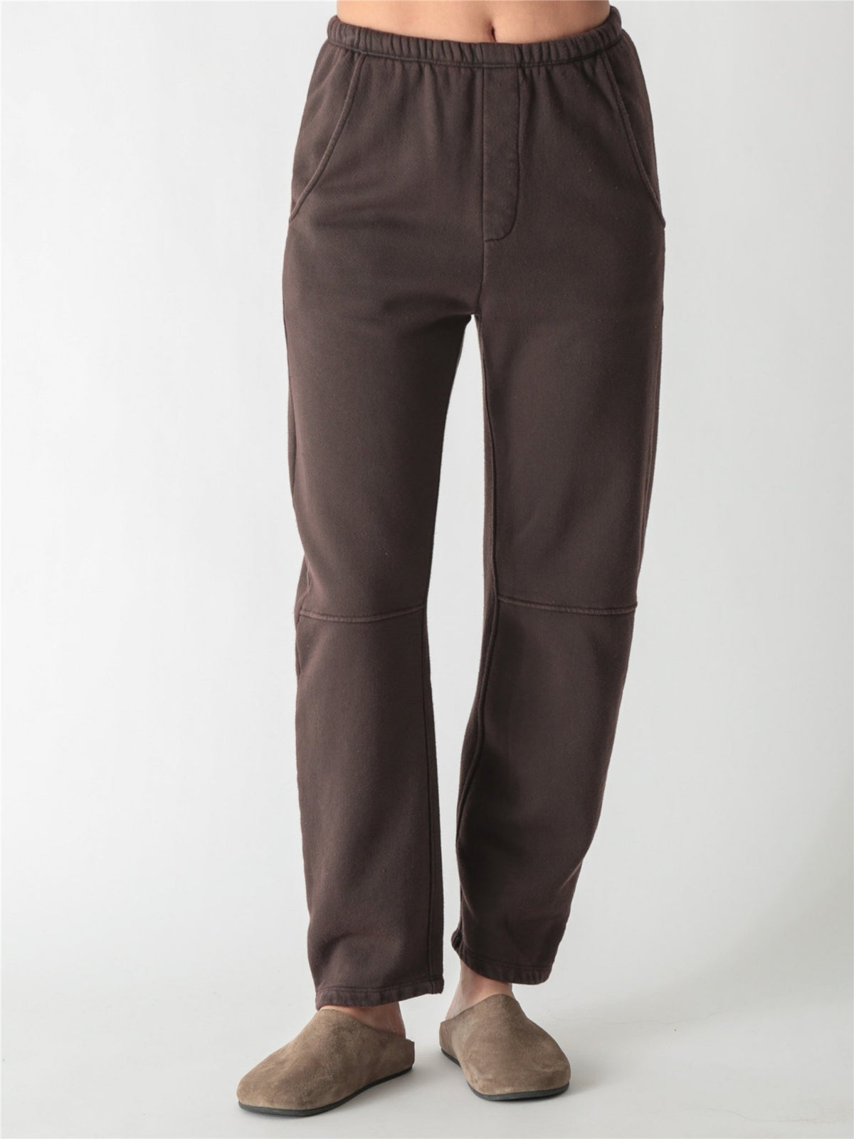 A person wearing dark brown relaxed-fit fleece sweatpants with elastic waistband, side pockets, and knee seams, paired with tan suede slip-on shoes against a plain white background.