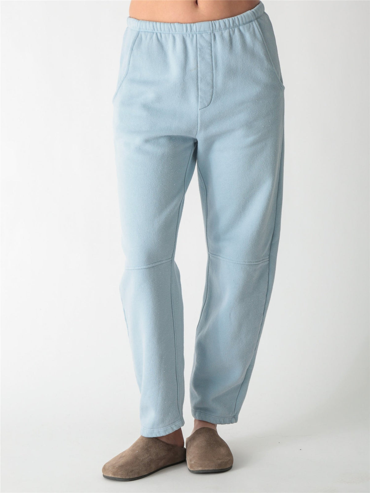 A person wearing ice blue barrel-fit fleece sweatpants with elastic waistband, side pockets, and knee seams, paired with tan suede slip-on shoes against a plain white background.