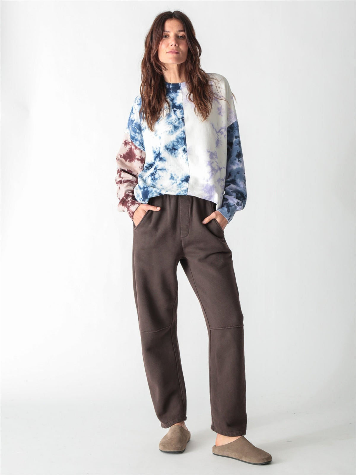 A person wearing dark brown relaxed-fit fleece sweatpants with elastic waistband, side pockets, and knee seams, paired with tan suede slip-on shoes against a plain white background.