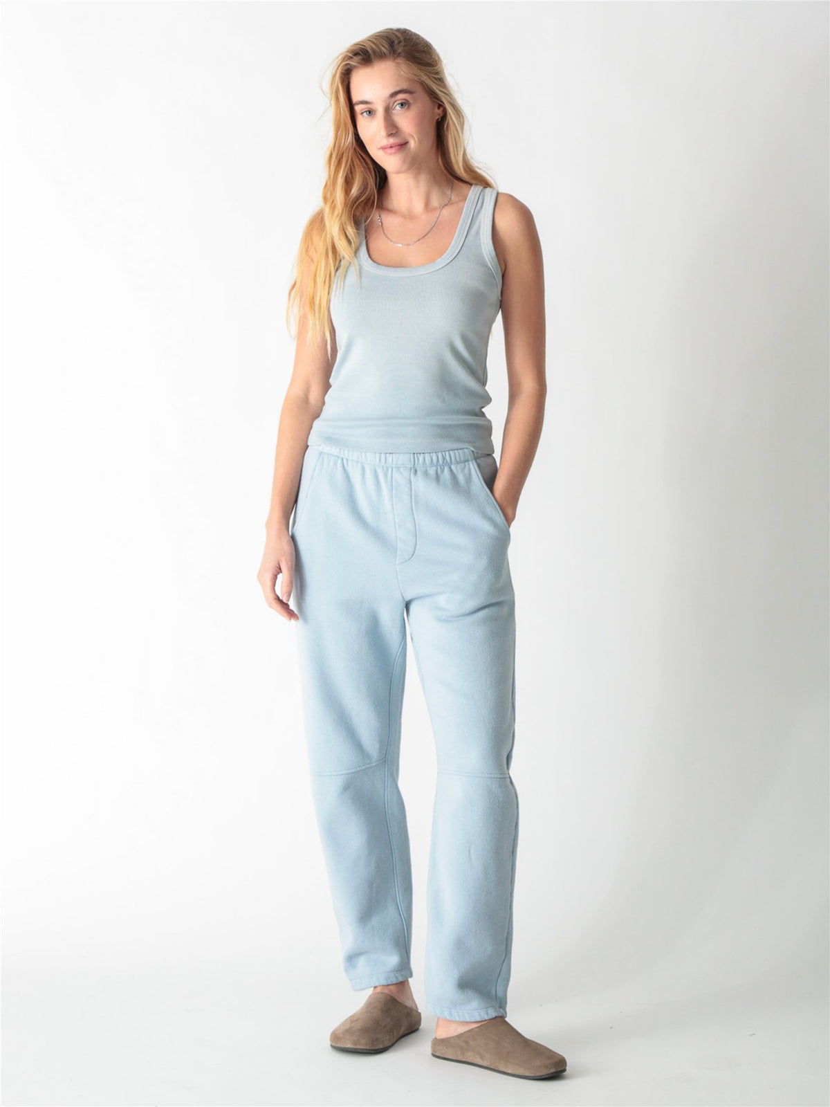 A person wearing ice blue barrel-fit fleece sweatpants with elastic waistband, side pockets, and knee seams, paired with tan suede slip-on shoes against a plain white background.