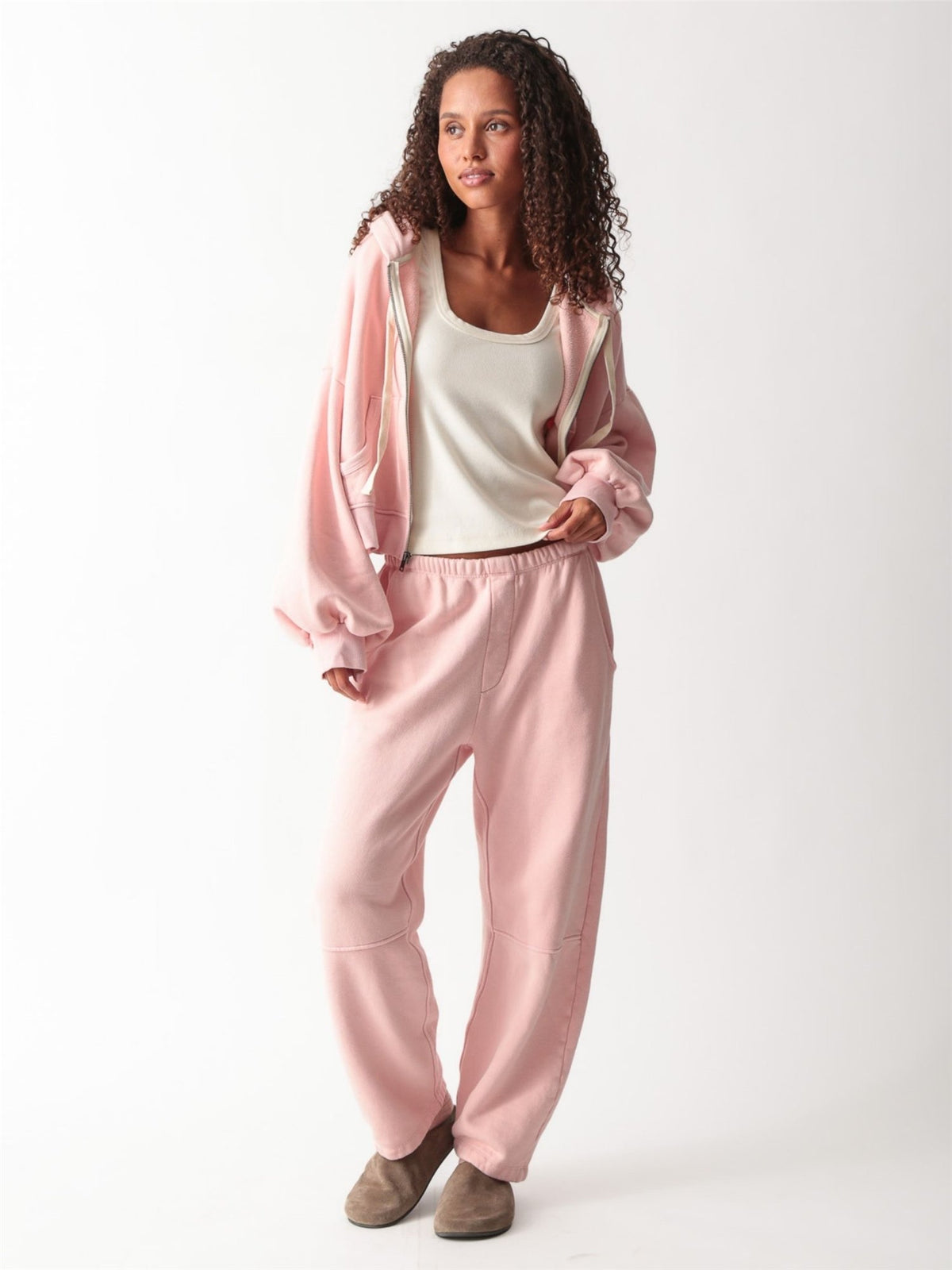 Model wearing a light pink cropped zip-up hoodie with balloon sleeves, open over an ivory scoop-neck tank, with matching pink barrel-leg sweatpants and brown suede clogs.