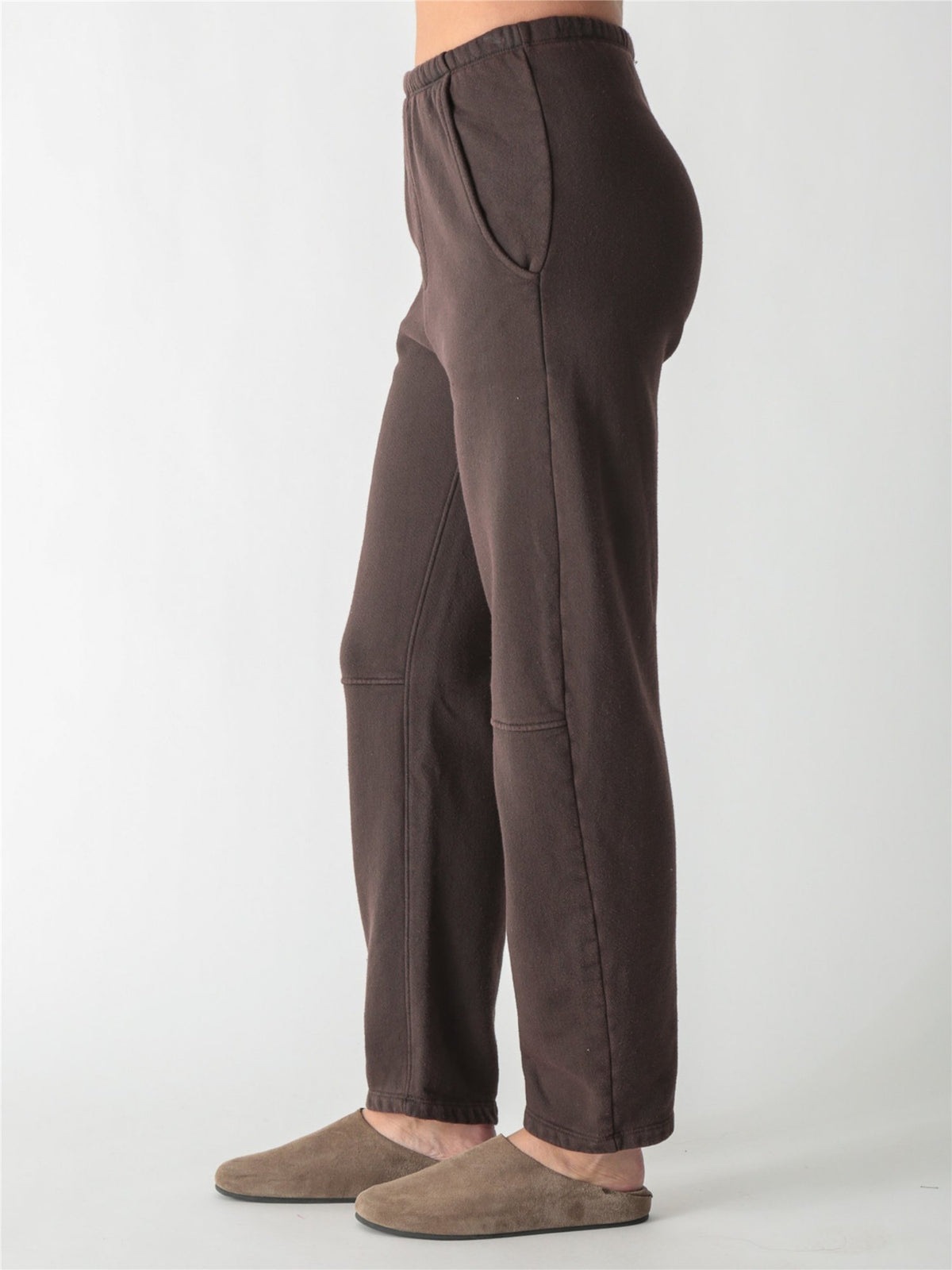 A person wearing dark brown relaxed-fit fleece sweatpants with elastic waistband, side pockets, and knee seams, paired with tan suede slip-on shoes against a plain white background.