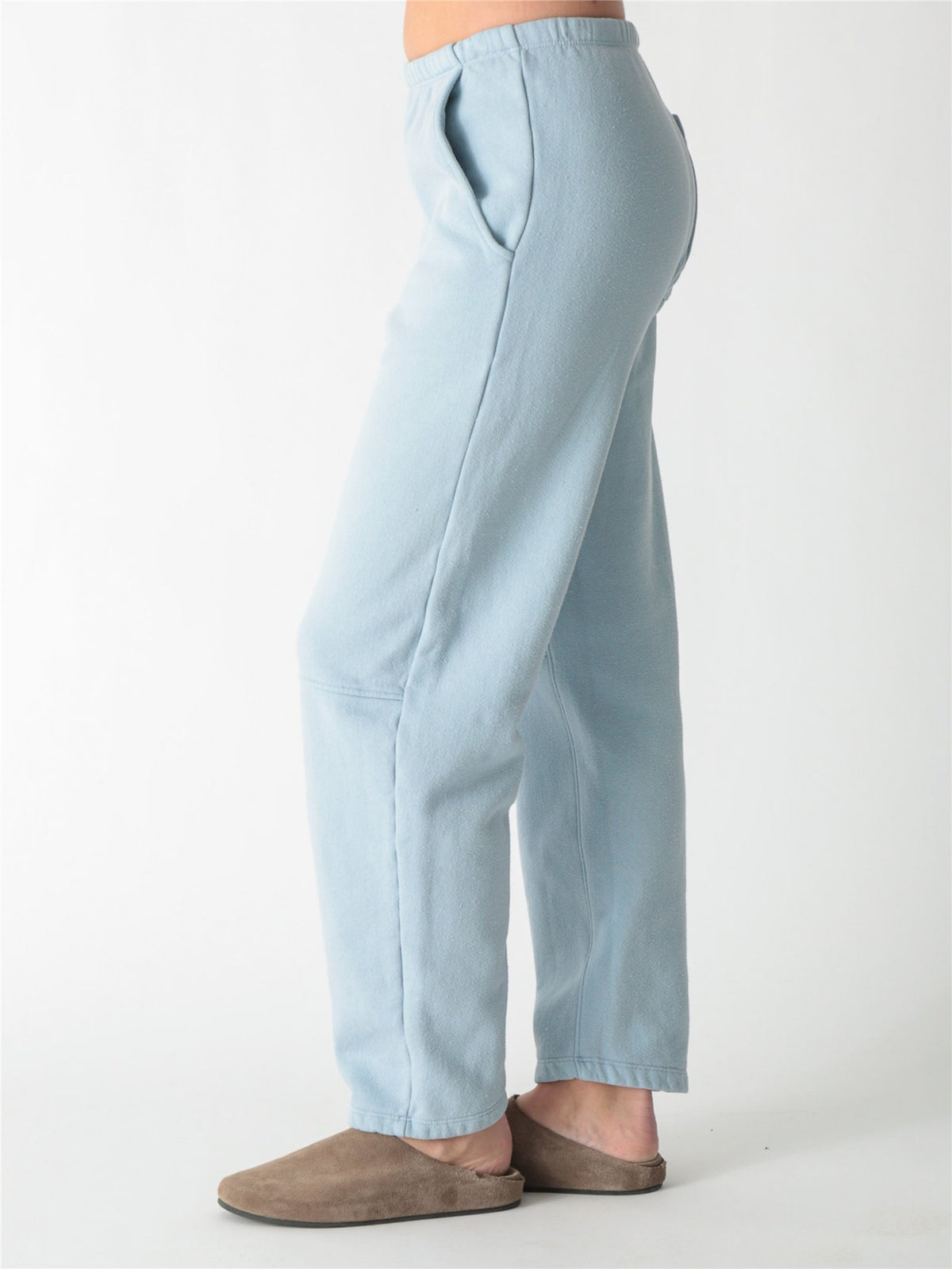 A person wearing ice blue barrel-fit fleece sweatpants with elastic waistband, side pockets, and knee seams, paired with tan suede slip-on shoes against a plain white background.