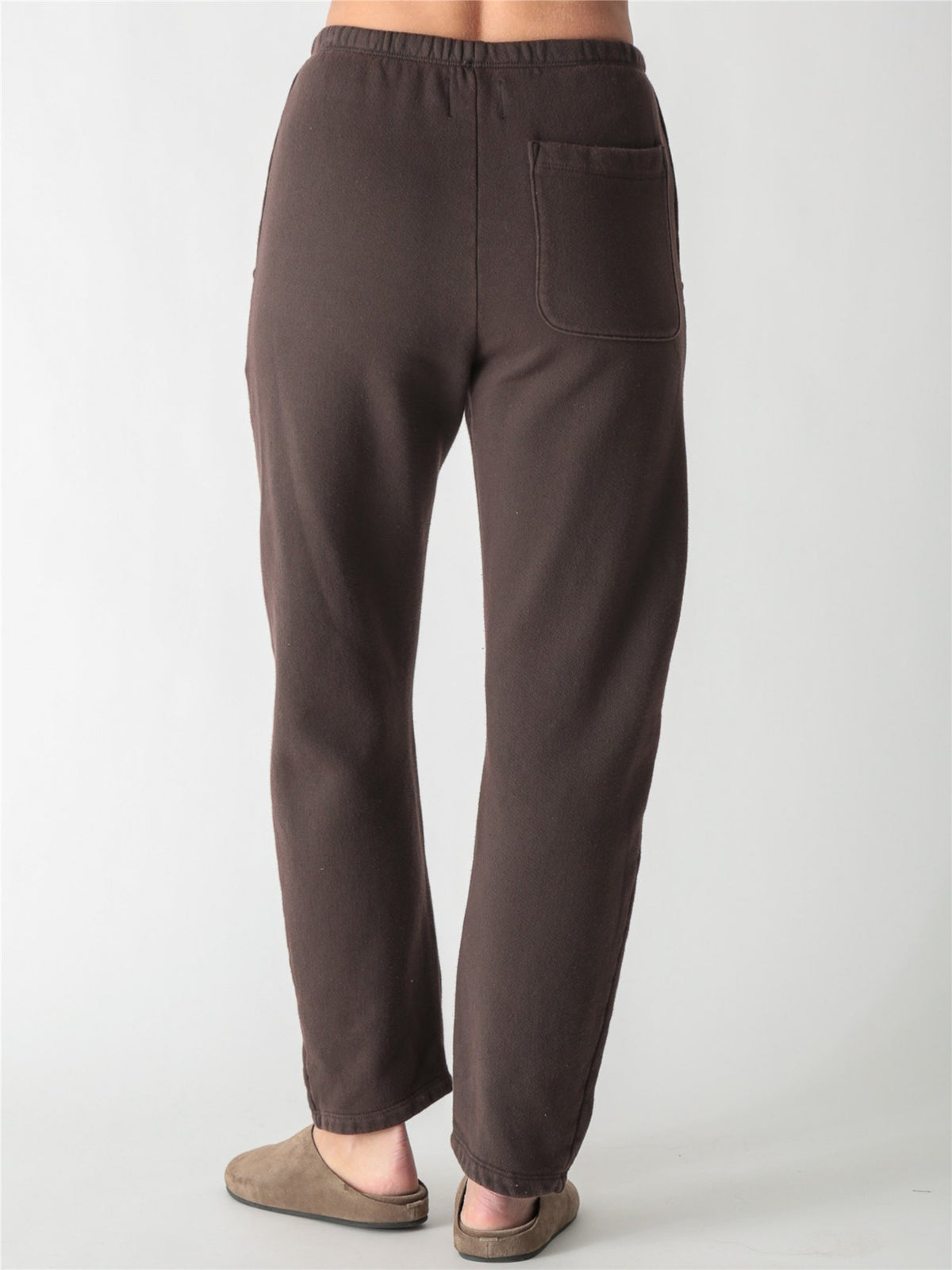 A person wearing dark brown relaxed-fit fleece sweatpants with elastic waistband, side pockets, and knee seams, paired with tan suede slip-on shoes against a plain white background.