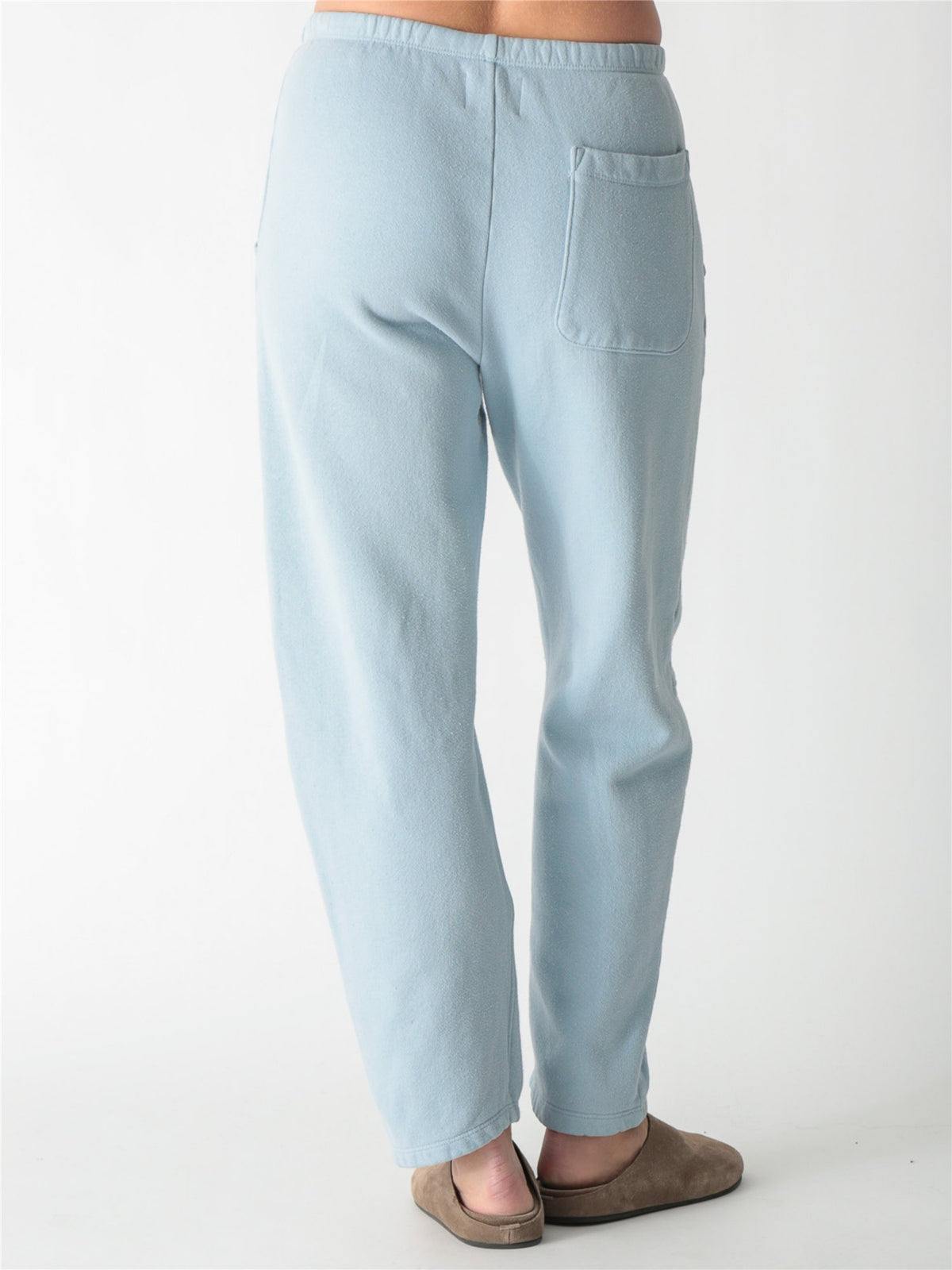 A person wearing ice blue barrel-fit fleece sweatpants with elastic waistband, side pockets, and knee seams, paired with tan suede slip-on shoes against a plain white background.