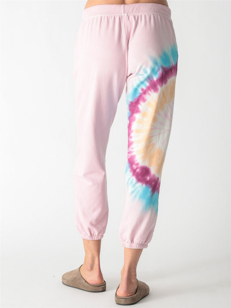 Light pink sweatpants featuring a drawstring waist, side pockets, elastic cuffs, and a vibrant tie-dye accent panel along the right leg.