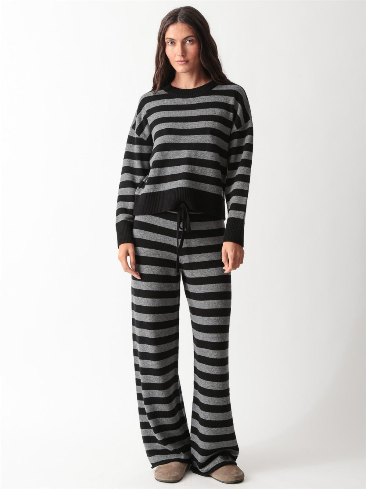 Woman wearing a black and heather grey striped knit crewneck sweater with matching wide-leg drawstring knit pants and slides.
