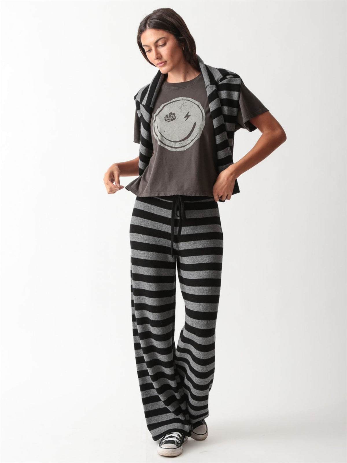 Woman wearing a black and heather grey striped knit crewneck sweater and graphic tee with matching wide-leg drawstring knit pants and white sneakers.