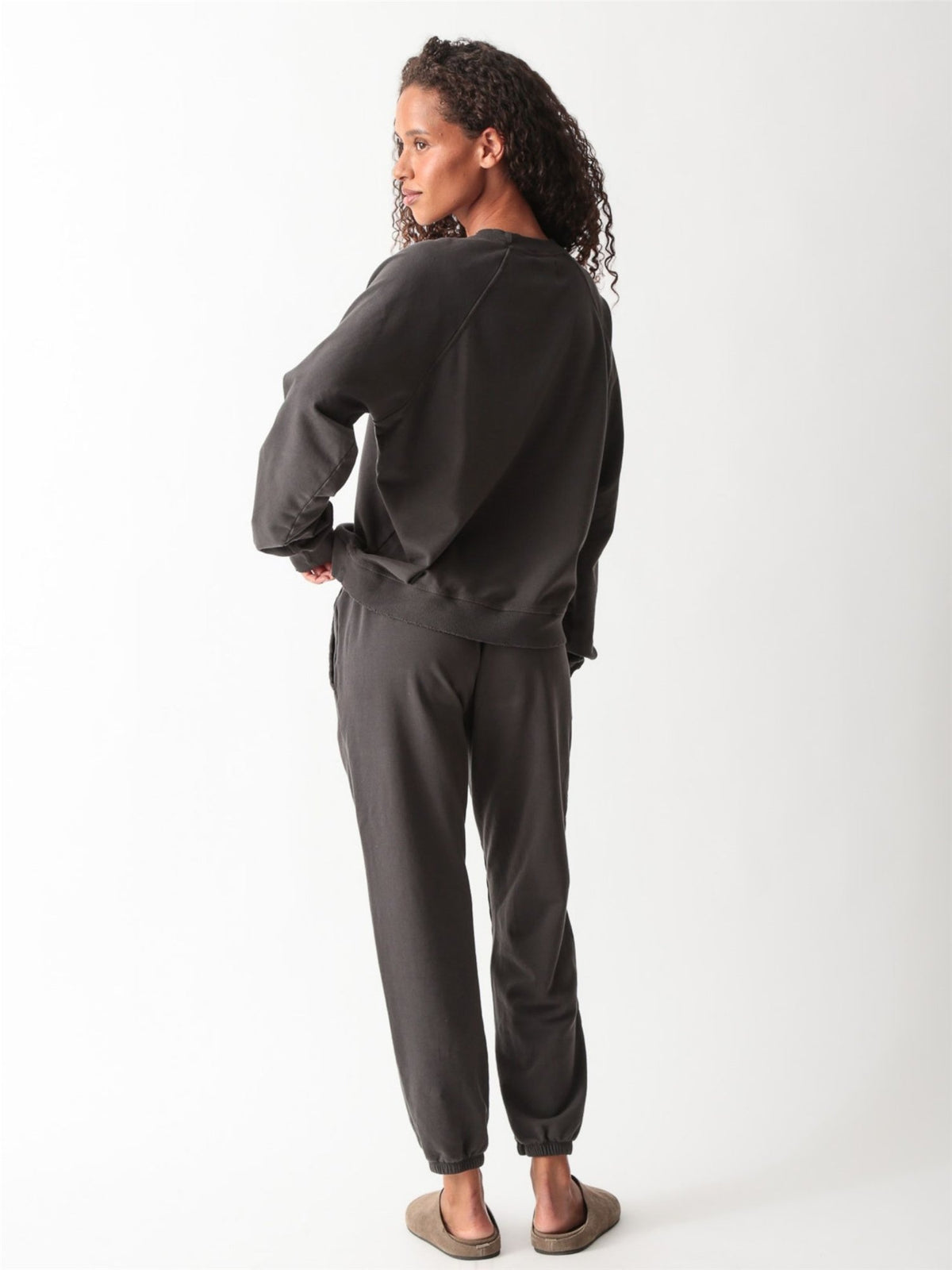 Woman wearing a washed charcoal raglan crewneck sweatshirt with matching drawstring jogger sweatpants and suede clogs.