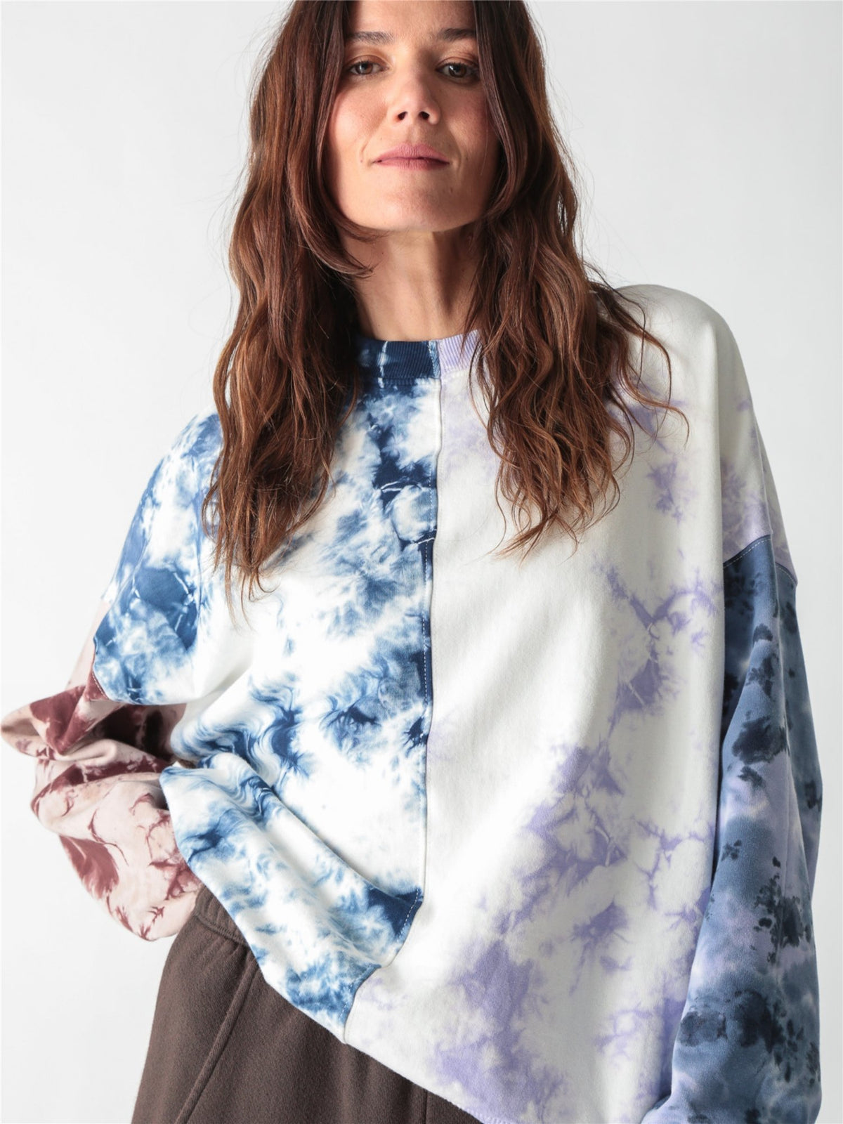 A woman wearing an oversized, multi-color tie-dye sweatshirt featuring split panels of blue, purple, and brown tie-dye patterns set against a white background.