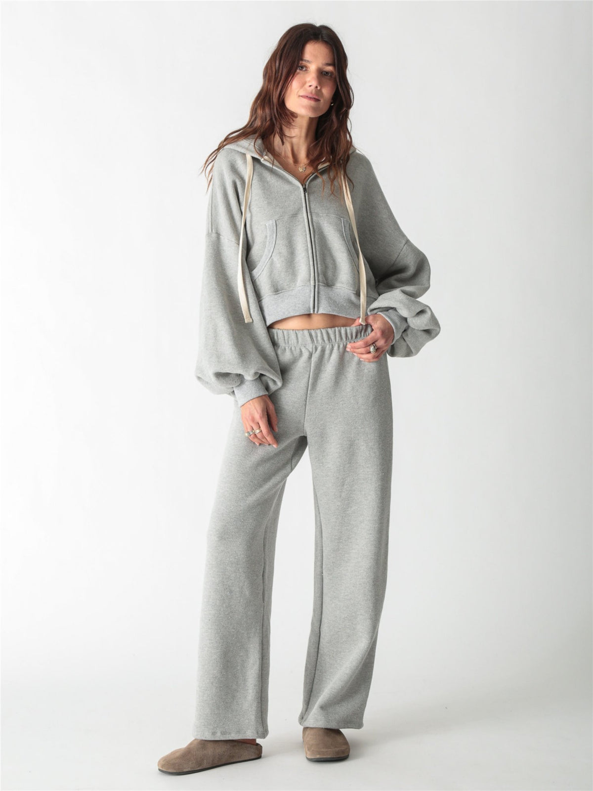 A woman wearing a heather gray cropped zip-up hoodie with voluminous sleeves and matching wide-leg sweatpants against a white background.