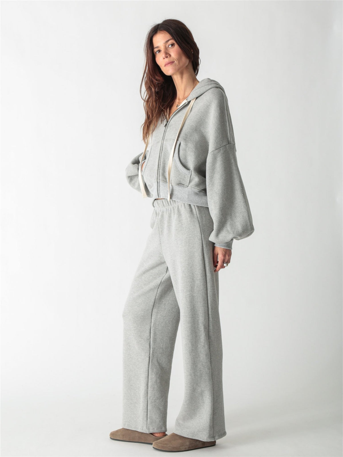 A woman wearing a heather gray cropped zip-up hoodie with voluminous sleeves and matching wide-leg sweatpants against a white background.