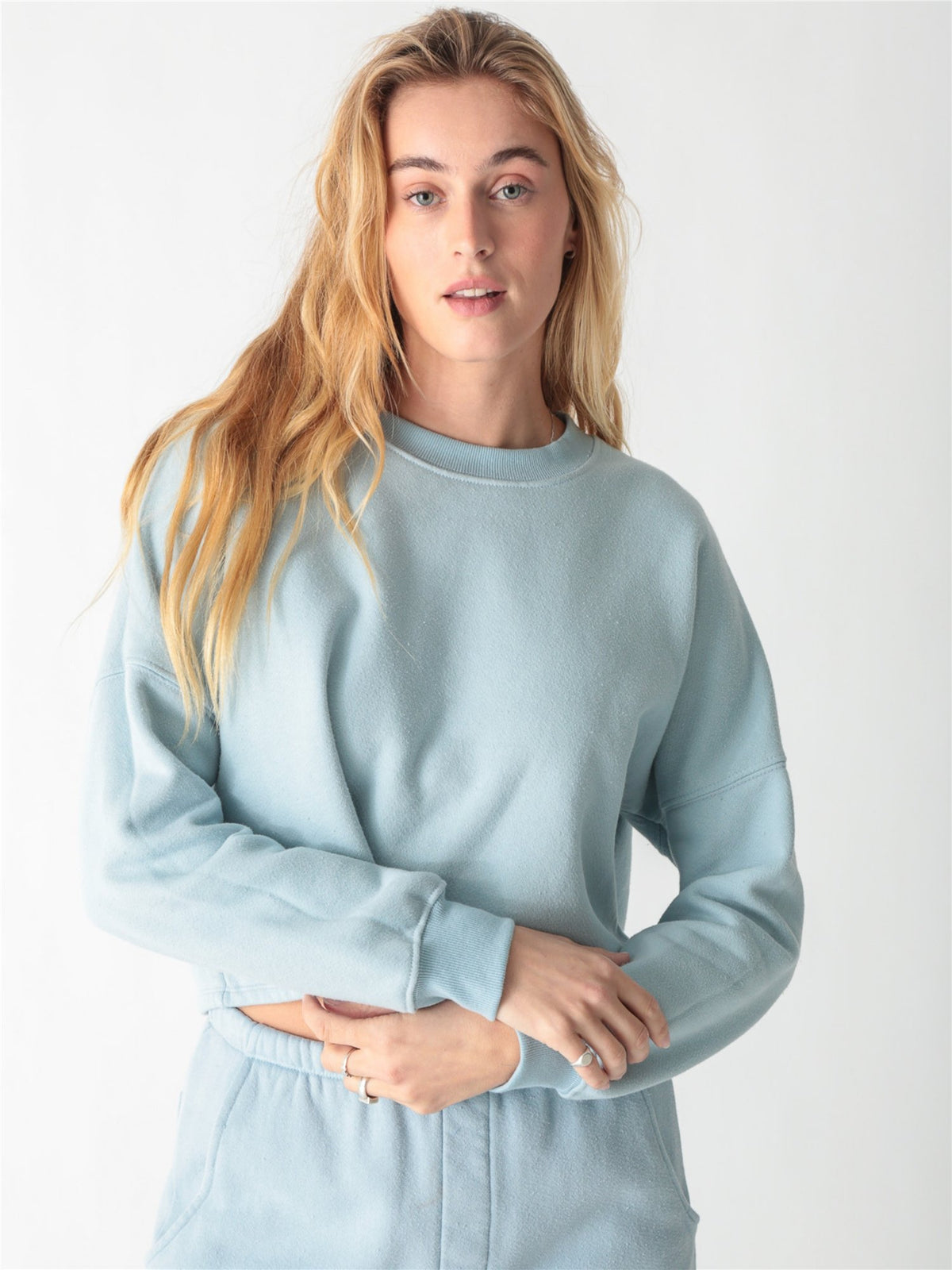A woman wearing a light blue crewneck sweatshirt with ribbed cuffs and matching sweatpants against a white background.