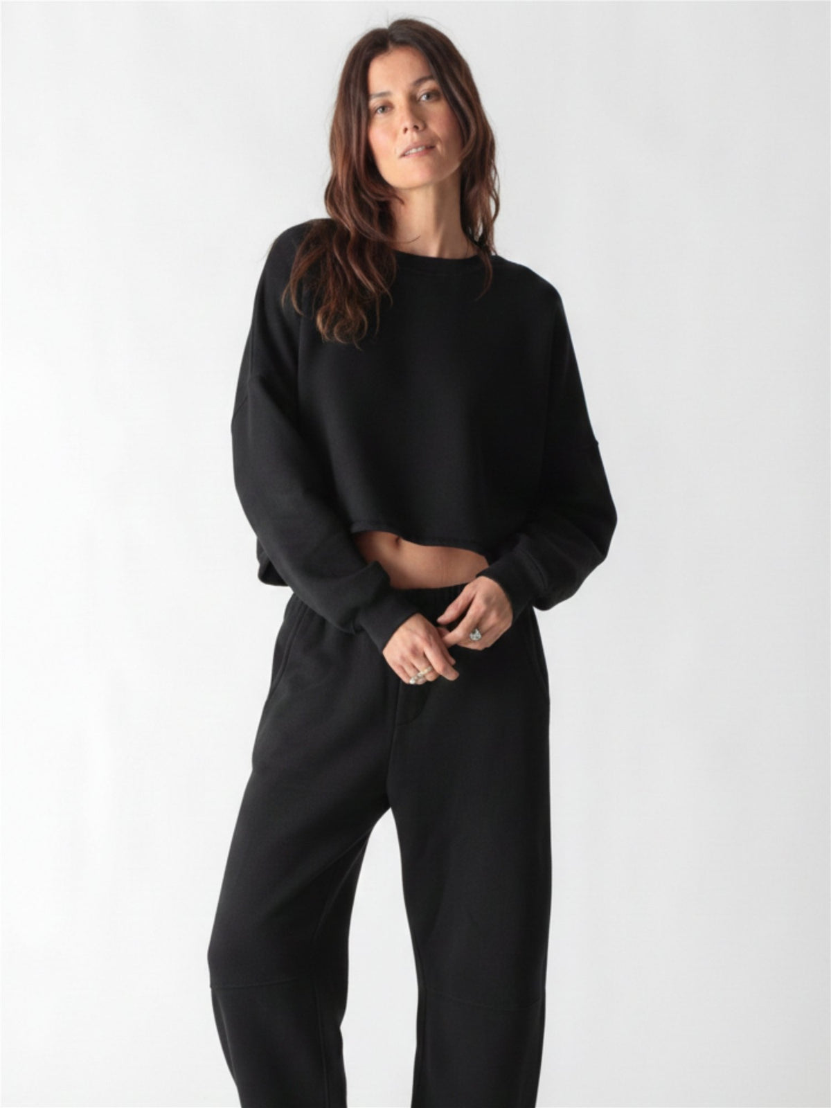A woman wearing a black crewneck sweatshirt with ribbed cuffs and matching sweatpants against a white background.