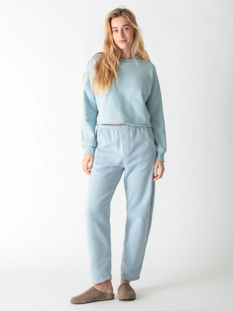 A woman wearing a light blue crewneck sweatshirt with ribbed cuffs and matching sweatpants against a white background.