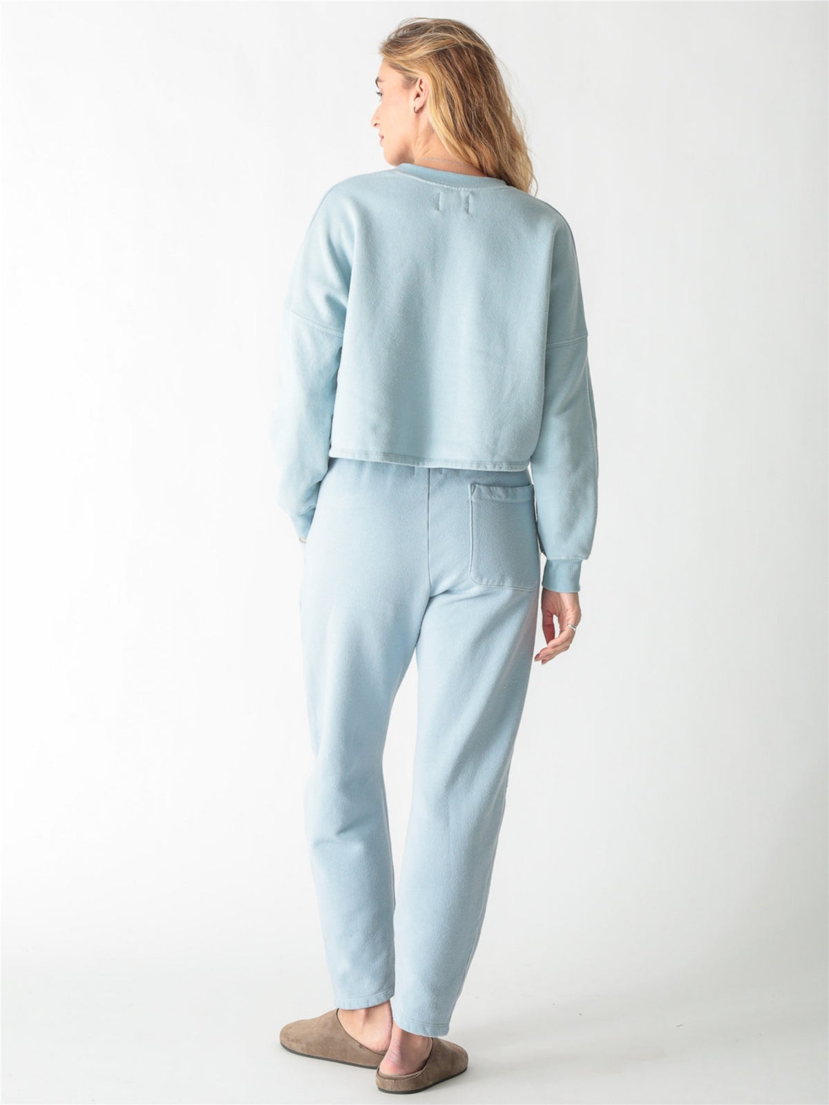 A woman wearing a light blue crewneck sweatshirt with ribbed cuffs and matching sweatpants against a white background.