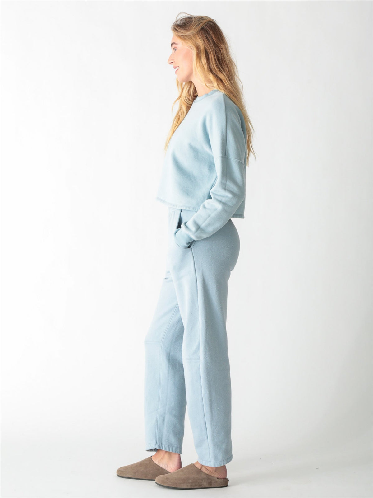 A woman wearing a light blue crewneck sweatshirt with ribbed cuffs and matching sweatpants against a white background.