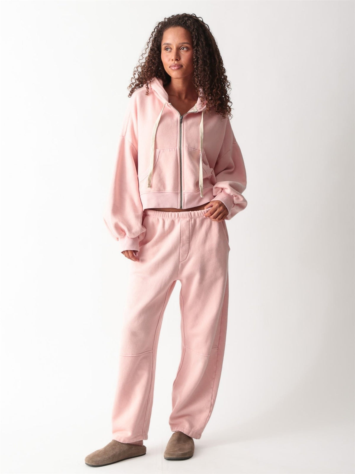 Model wearing a light pink cropped zip-up hoodie with balloon sleeves, open over an ivory scoop-neck tank, with matching pink barrel-leg sweatpants and brown suede clogs.