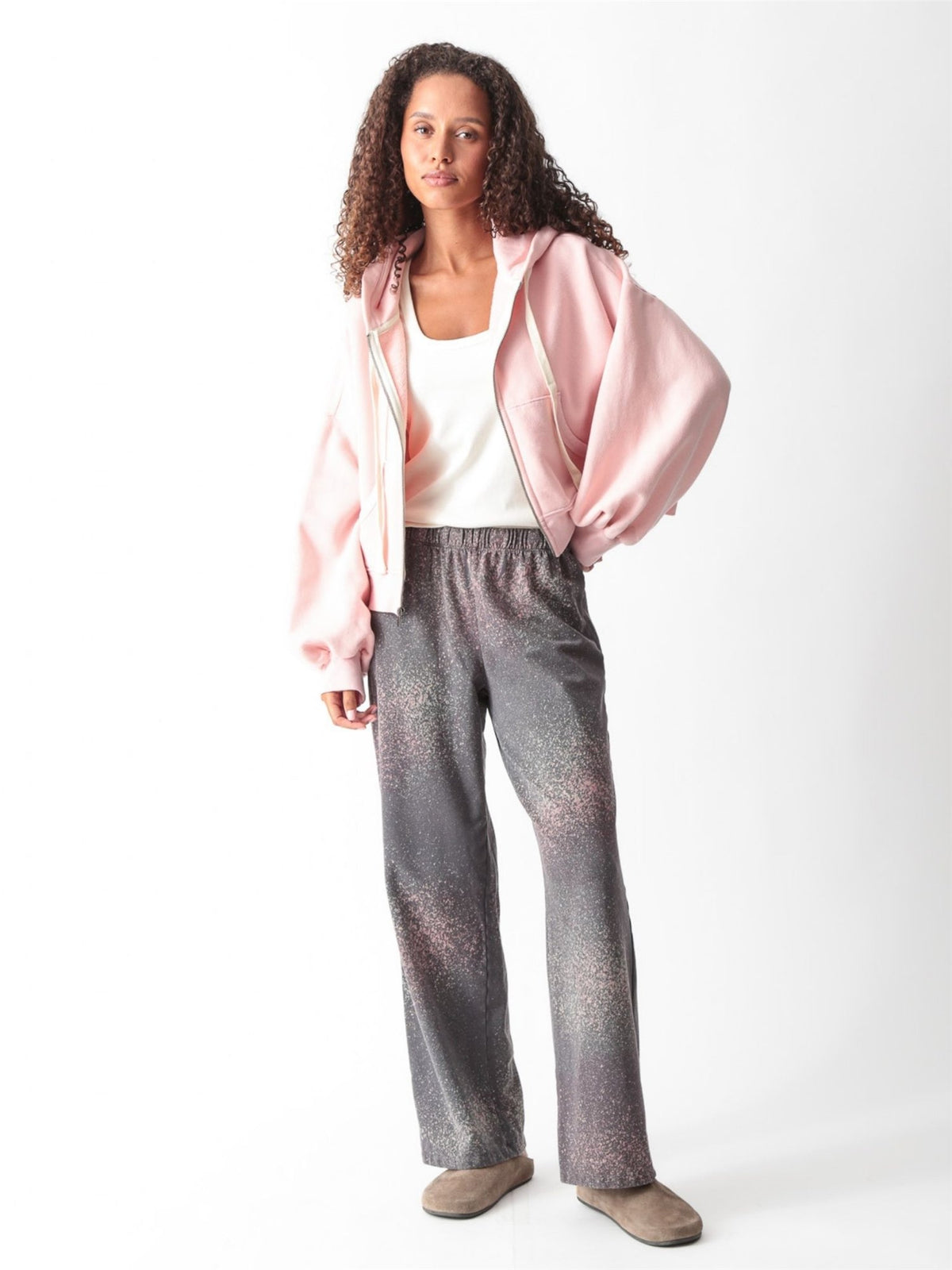 Model wearing a light pink cropped zip-up hoodie with balloon sleeves, open over an ivory scoop-neck tank, with matching pink barrel-leg sweatpants and brown suede clogs.