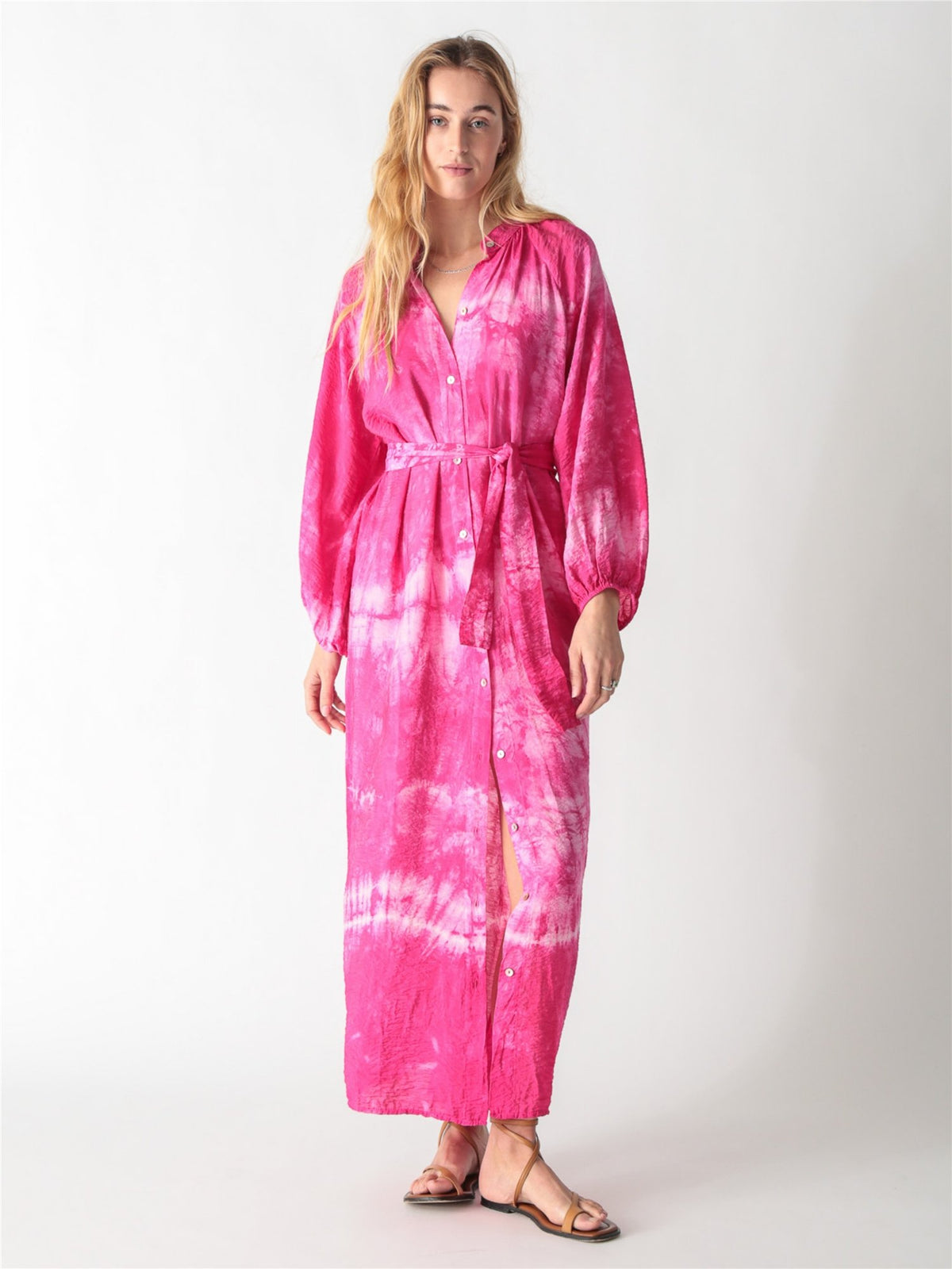 A woman wearing a vibrant pink tie-dye maxi shirt dress with long billowy sleeves, a button-down front, and a tied waist belt.