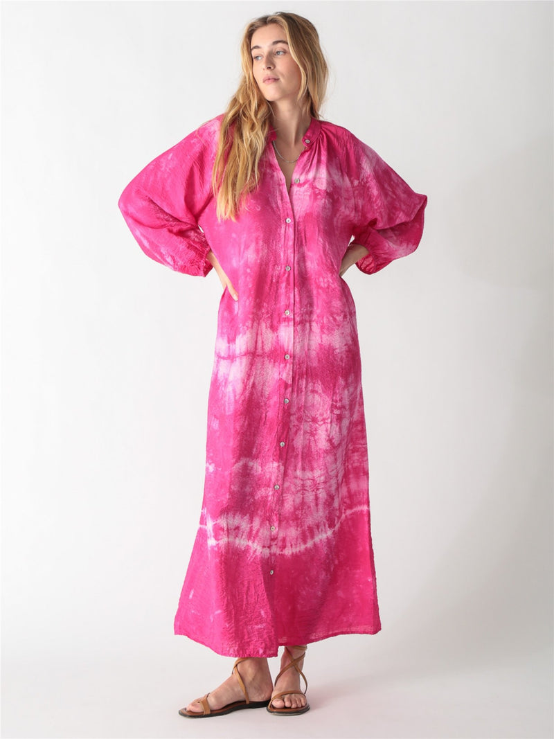 A woman wearing a vibrant pink tie-dye maxi shirt dress with long billowy sleeves, a button-down front, and a tied waist belt.