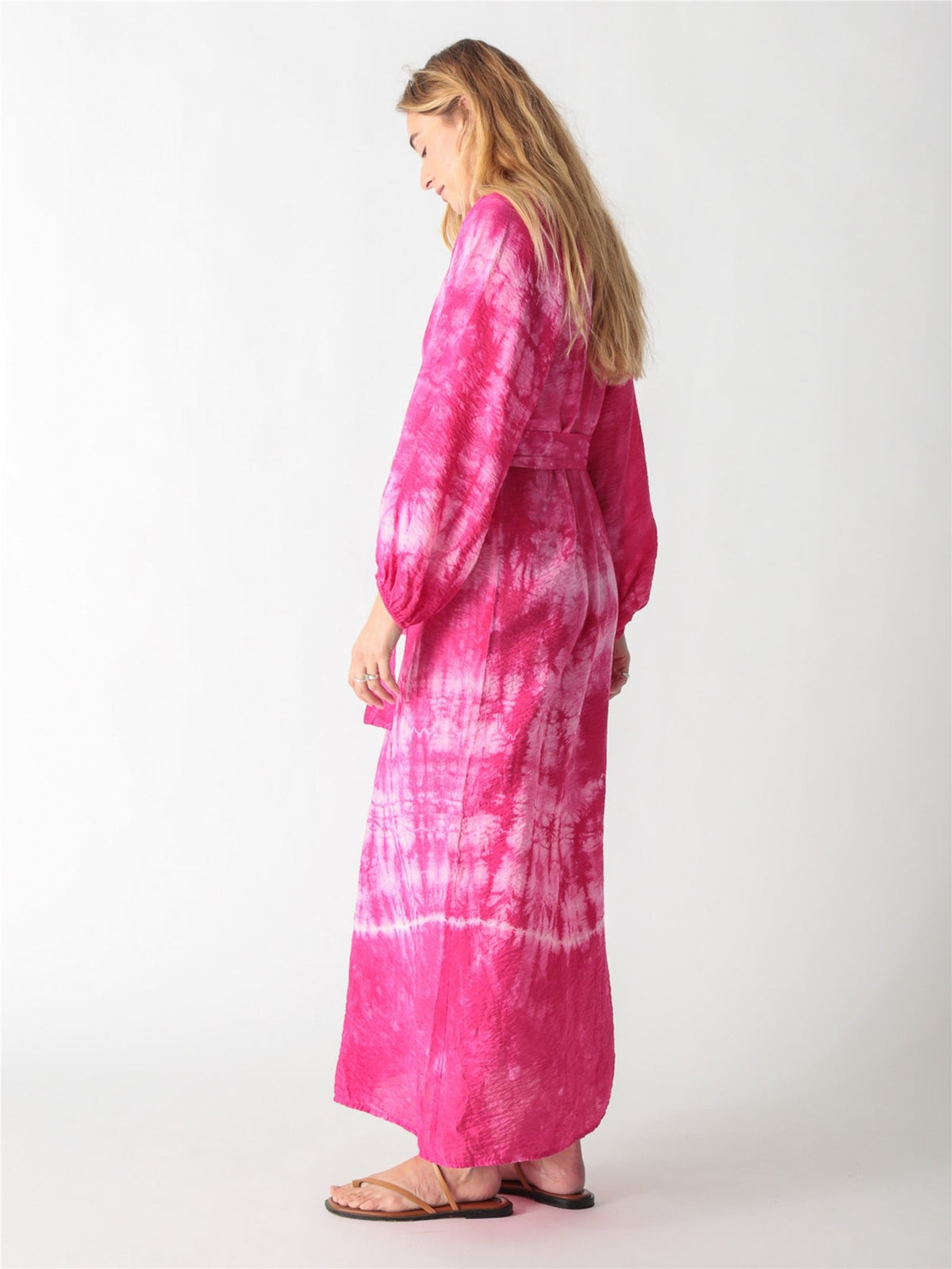 A woman wearing a vibrant pink tie-dye maxi shirt dress with long billowy sleeves, a button-down front, and a tied waist belt.