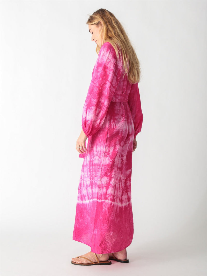 A woman wearing a vibrant pink tie-dye maxi shirt dress with long billowy sleeves, a button-down front, and a tied waist belt.