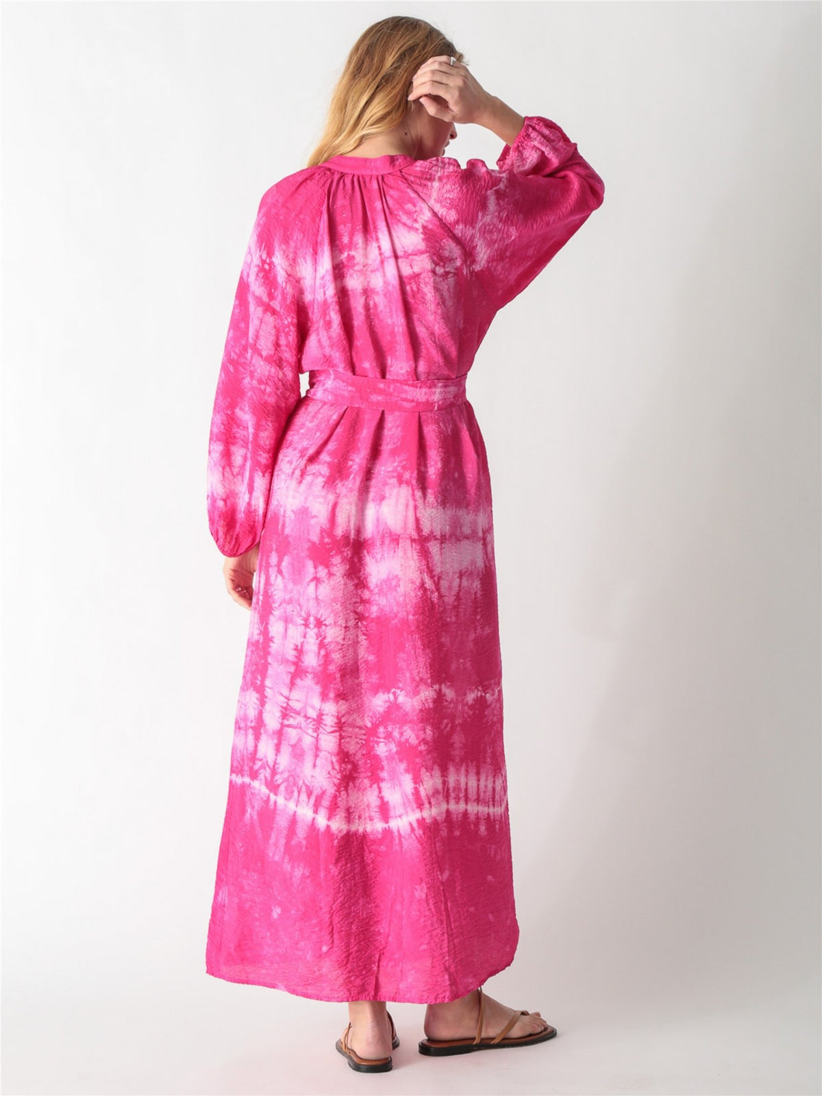 A woman wearing a vibrant pink tie-dye maxi shirt dress with long billowy sleeves, a button-down front, and a tied waist belt.