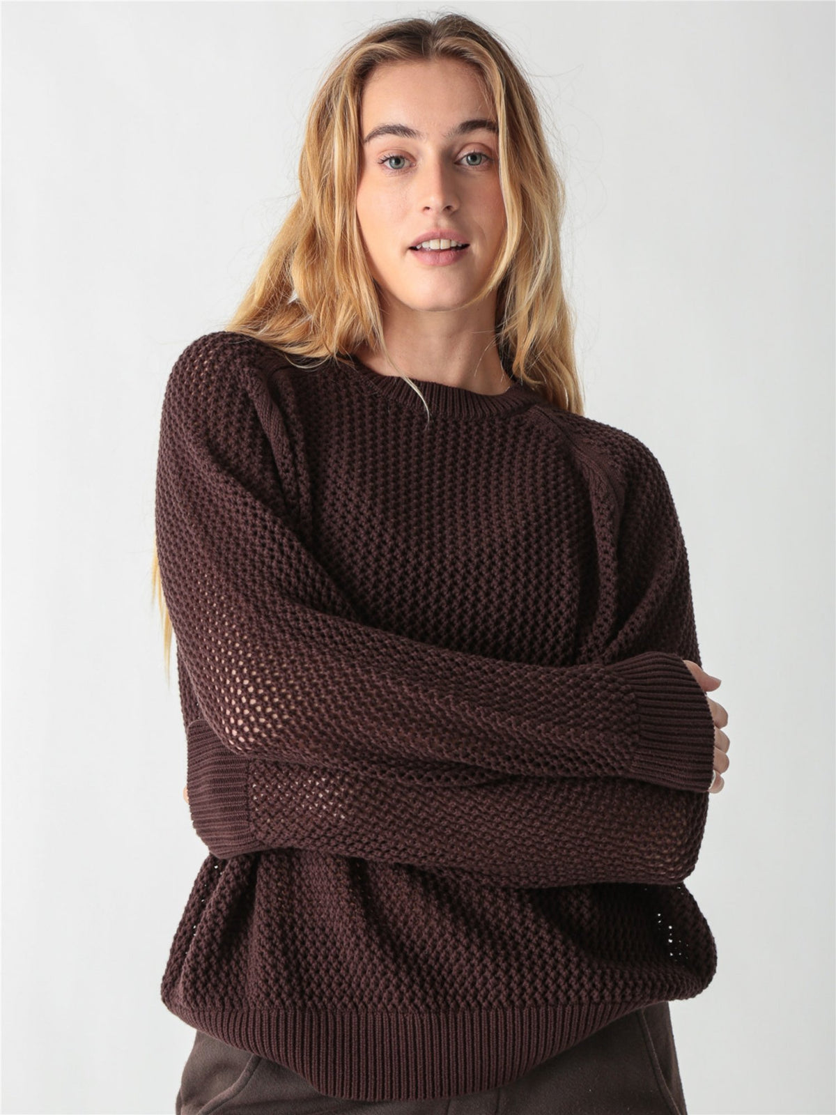 A woman with long blonde hair wearing a dark brown, chunky open-knit sweater with ribbed cuffs and hem against a plain white background.