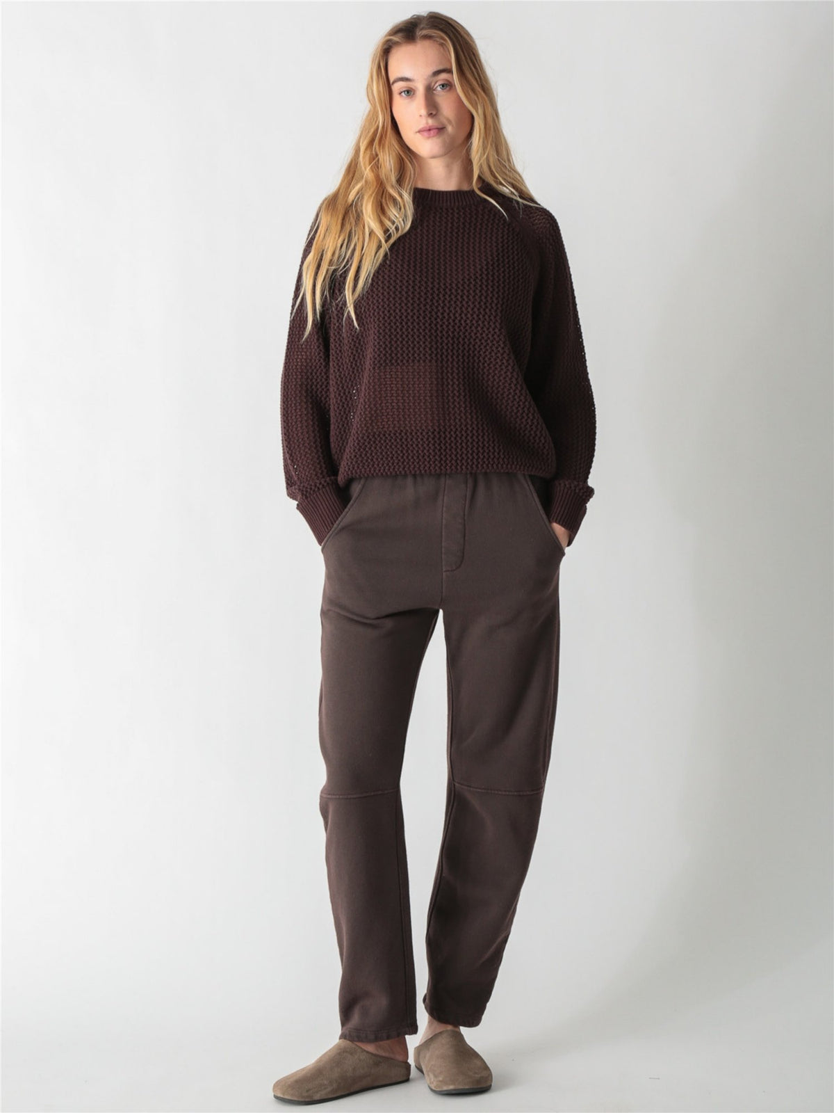 A woman with long blonde hair wearing a dark brown, chunky open-knit sweater with ribbed cuffs and hem against a plain white background.
