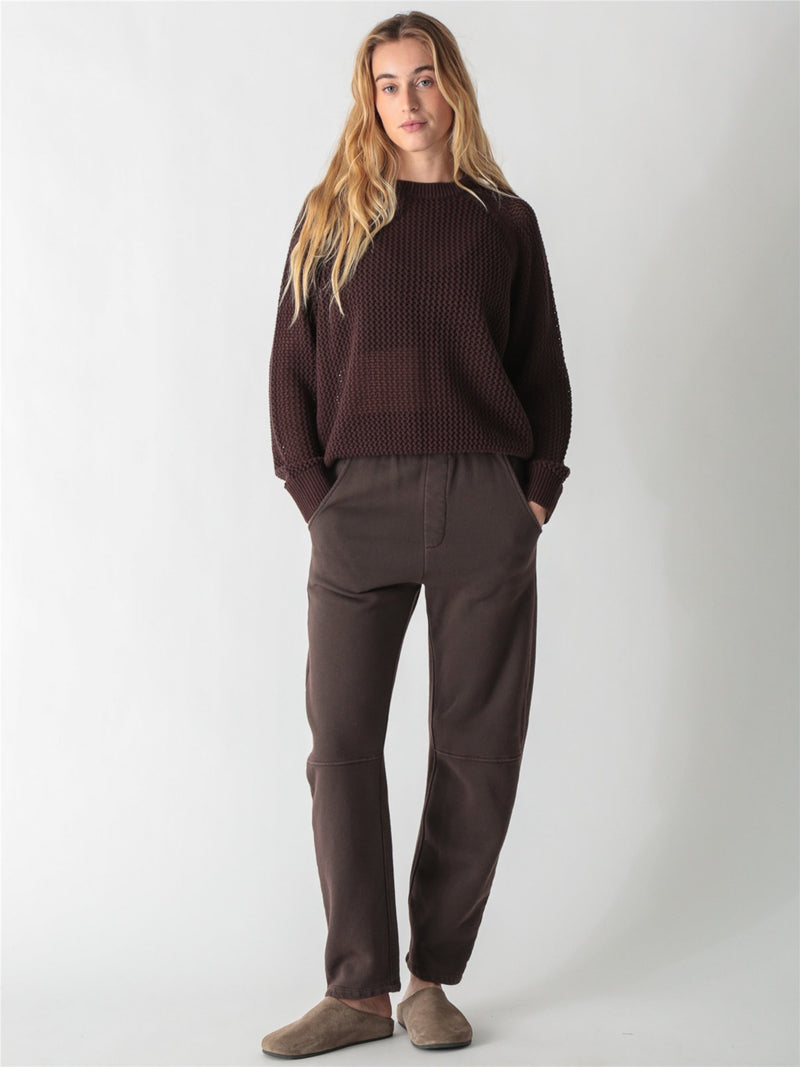 A woman with long blonde hair wearing a dark brown, chunky open-knit sweater with ribbed cuffs and hem against a plain white background.