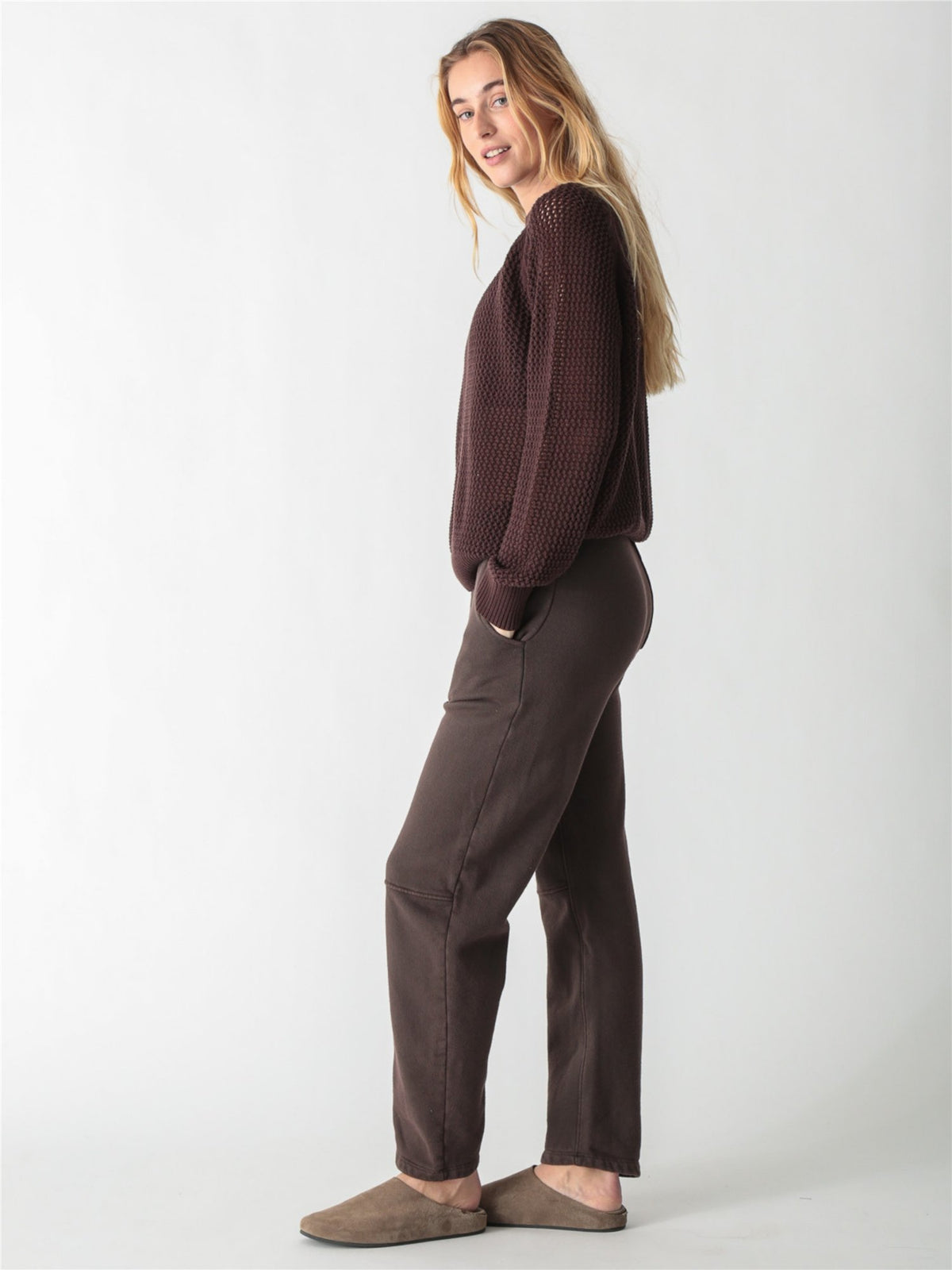 A woman with long blonde hair wearing a dark brown, chunky open-knit sweater with ribbed cuffs and hem against a plain white background.