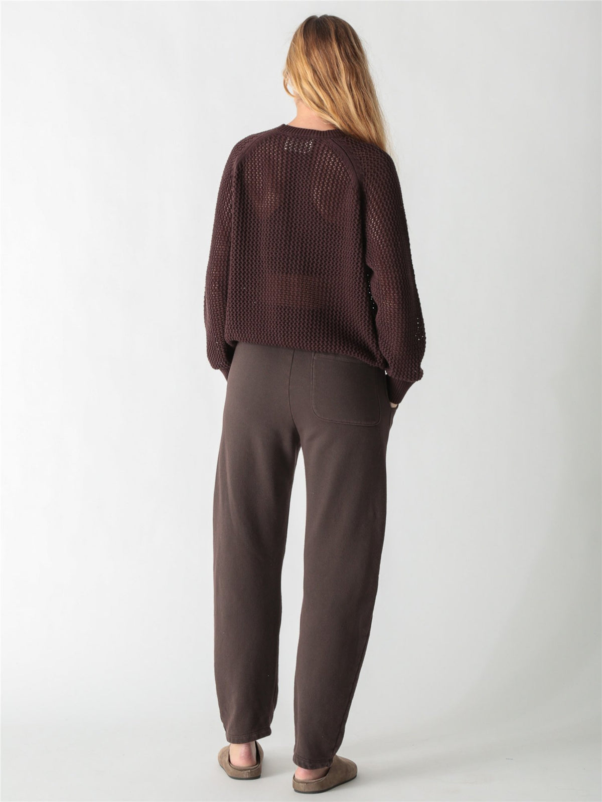 A woman with long blonde hair wearing a dark brown, chunky open-knit sweater with ribbed cuffs and hem against a plain white background.