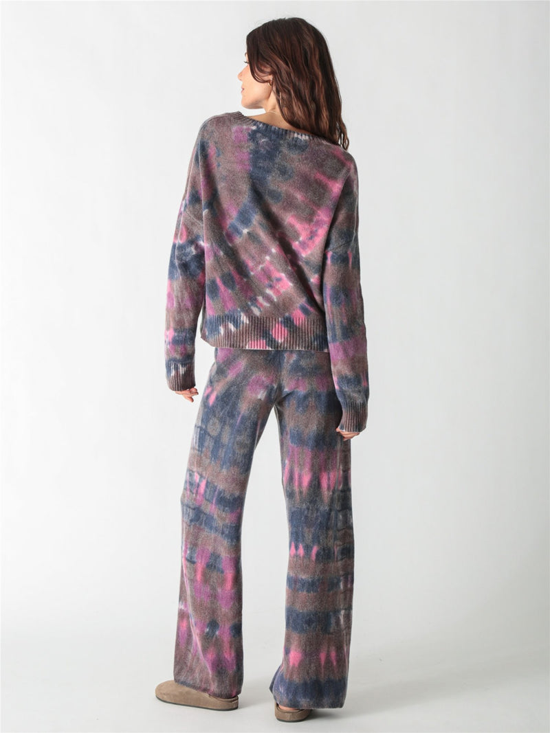 A long-sleeve, relaxed-fit knit sweater with a moody tie-dye pattern in shades of charcoal gray, muted purple, and bright pink accents.