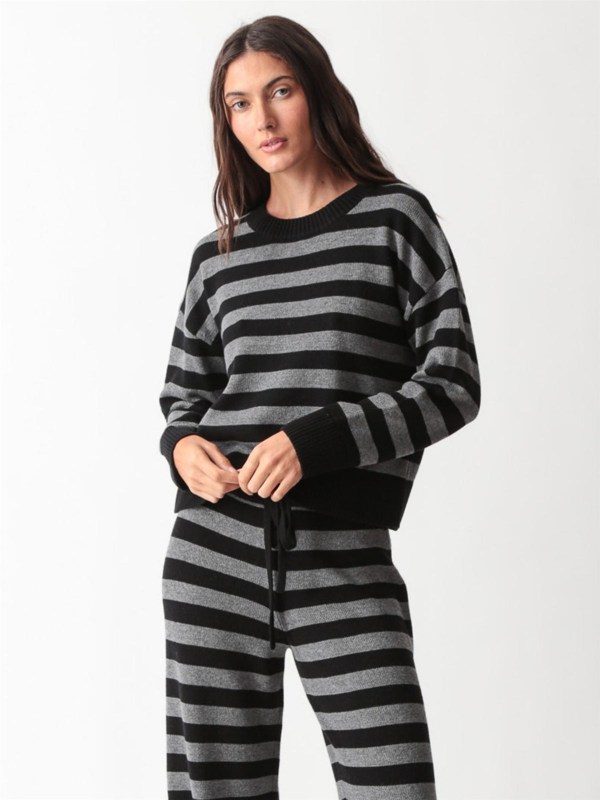 Woman wearing a black and heather grey striped knit crewneck sweater with matching wide-leg drawstring knit pants and white sneakers.