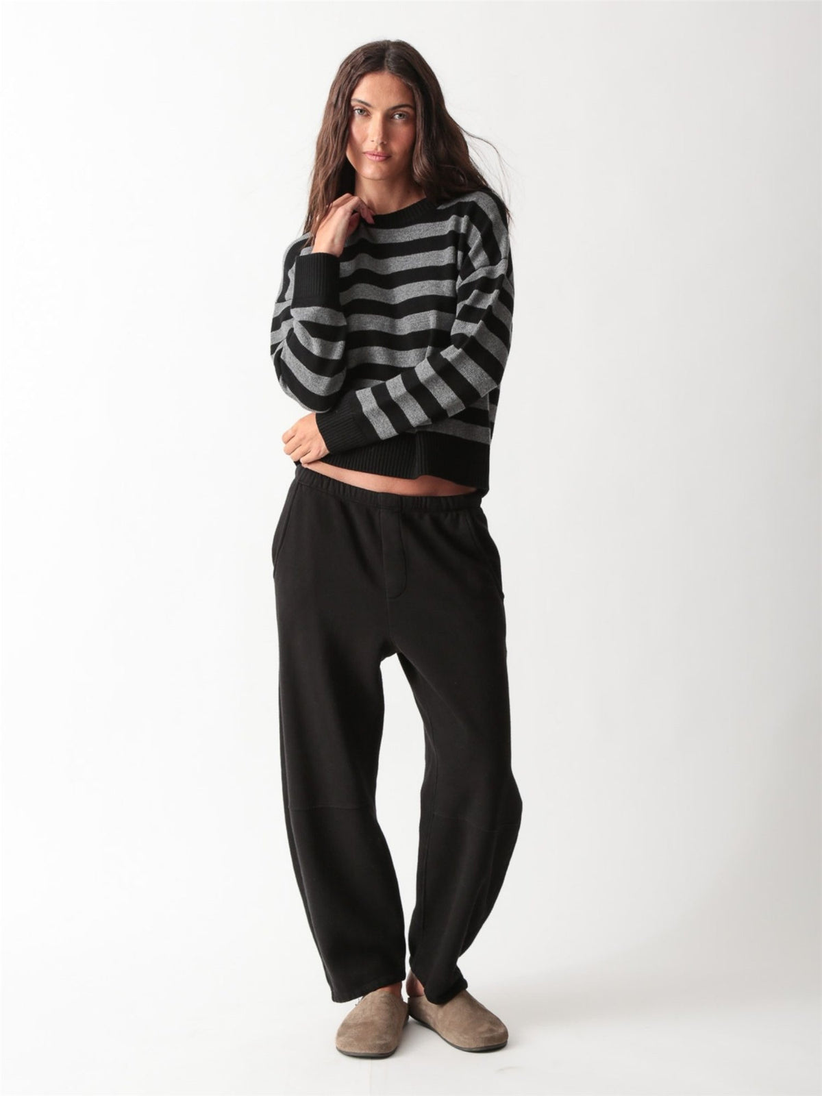 Woman wearing a black and heather grey striped knit crewneck sweater with black barrel leg sweatpants and slides.