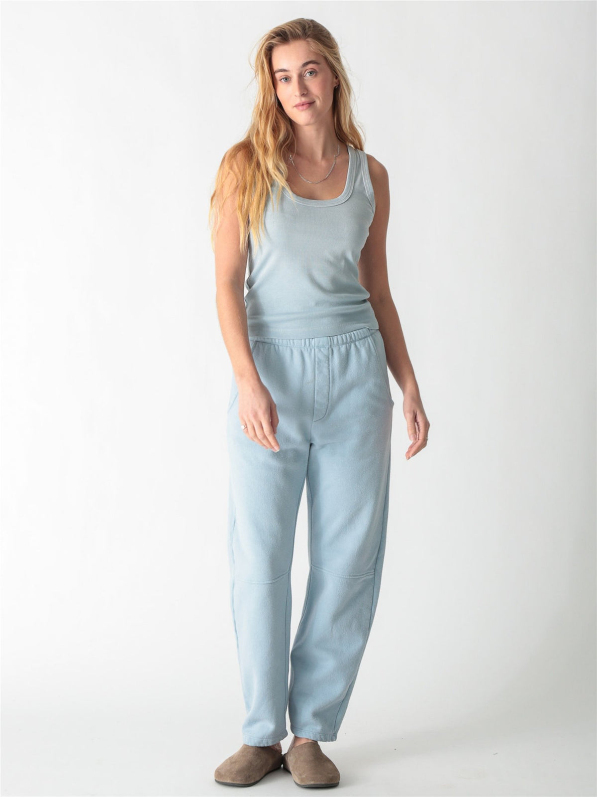 A woman wearing a light blue ribbed scoop neck tank top and matching sweatpants against a white background.