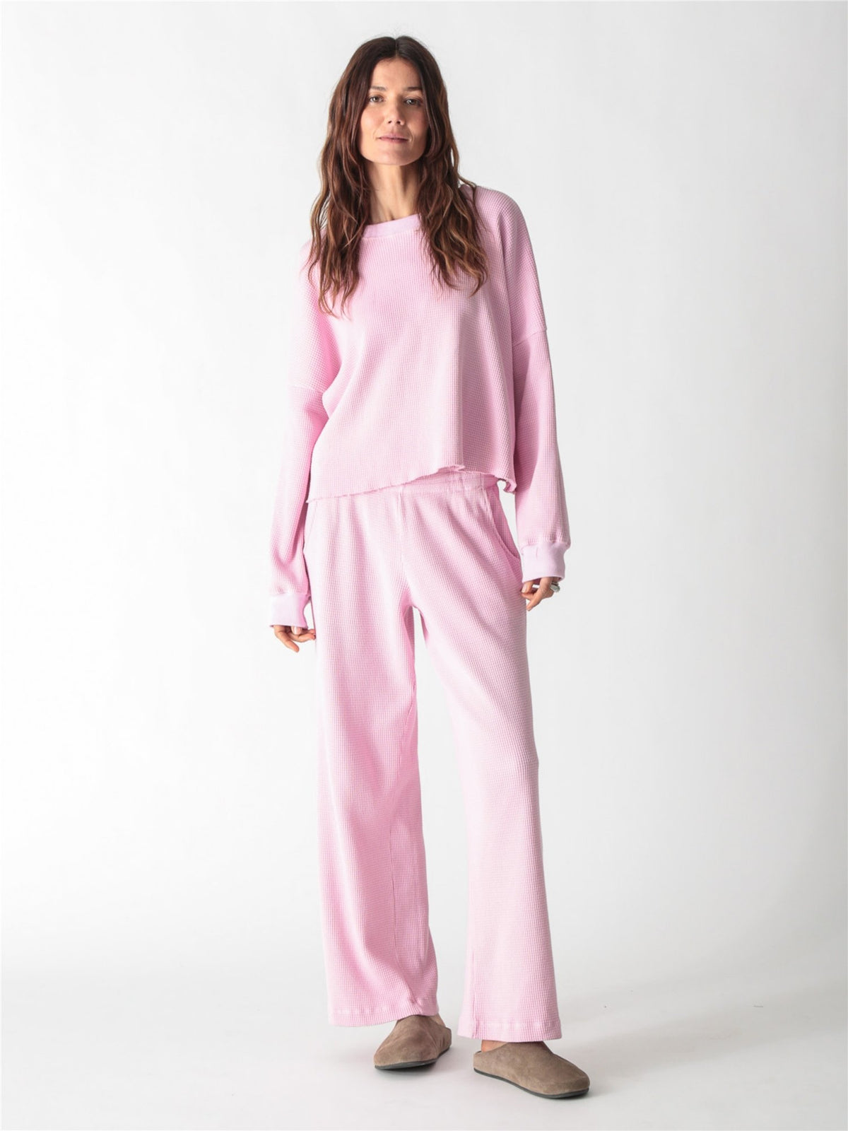 A woman wearing a light pink, long-sleeve waffle-knit lounge set with wide-leg pants against a plain white background.
