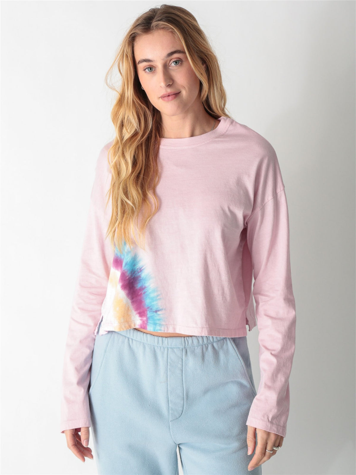 A woman wearing a light pink long-sleeve crop top featuring a colorful tie-dye accent along the right hem, paired with light blue sweatpants against a white background.